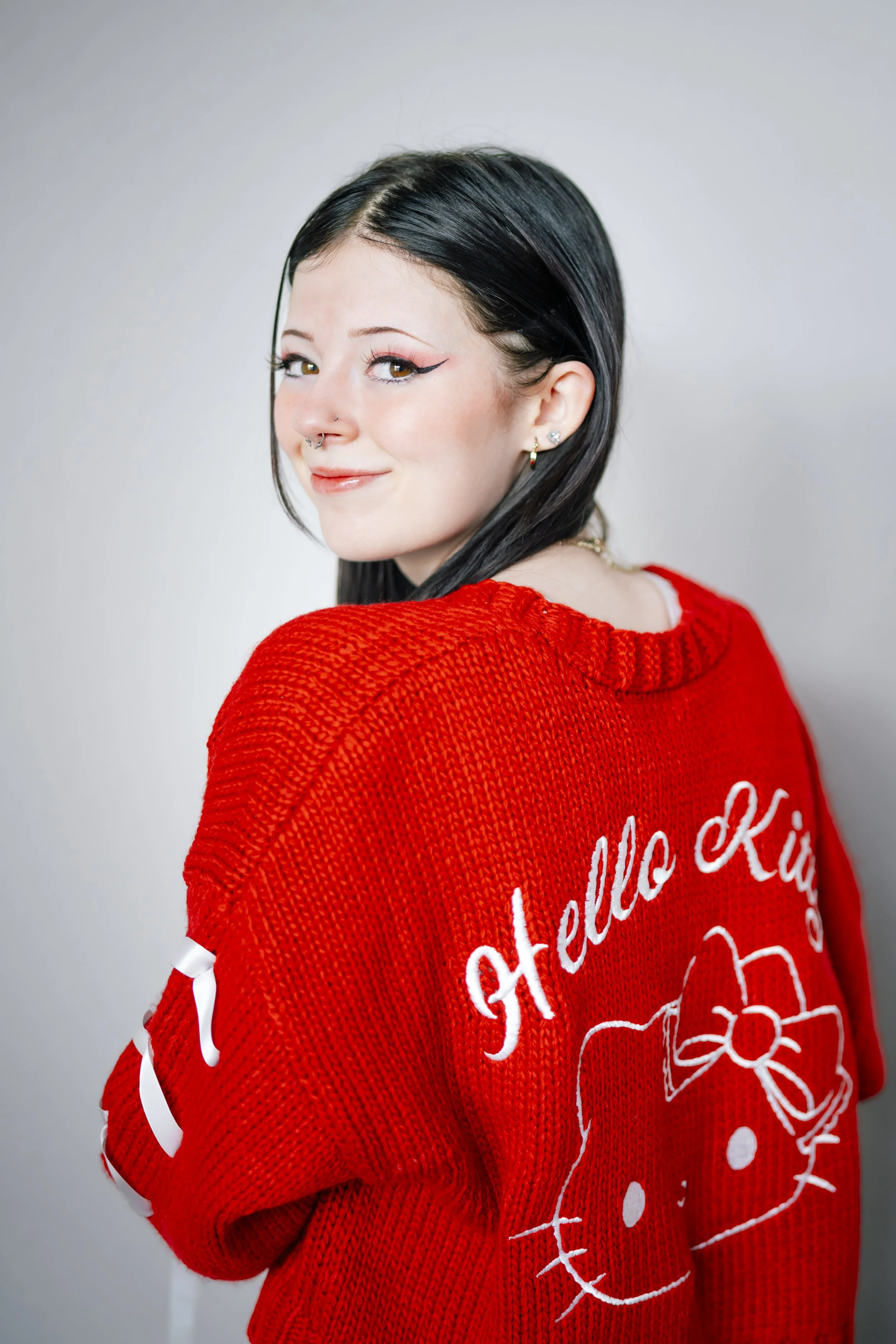 A young woman with black hair and multiple ear piercings is smiling and wearing a red Knit sweater with a Hello Kitty design on the back.
