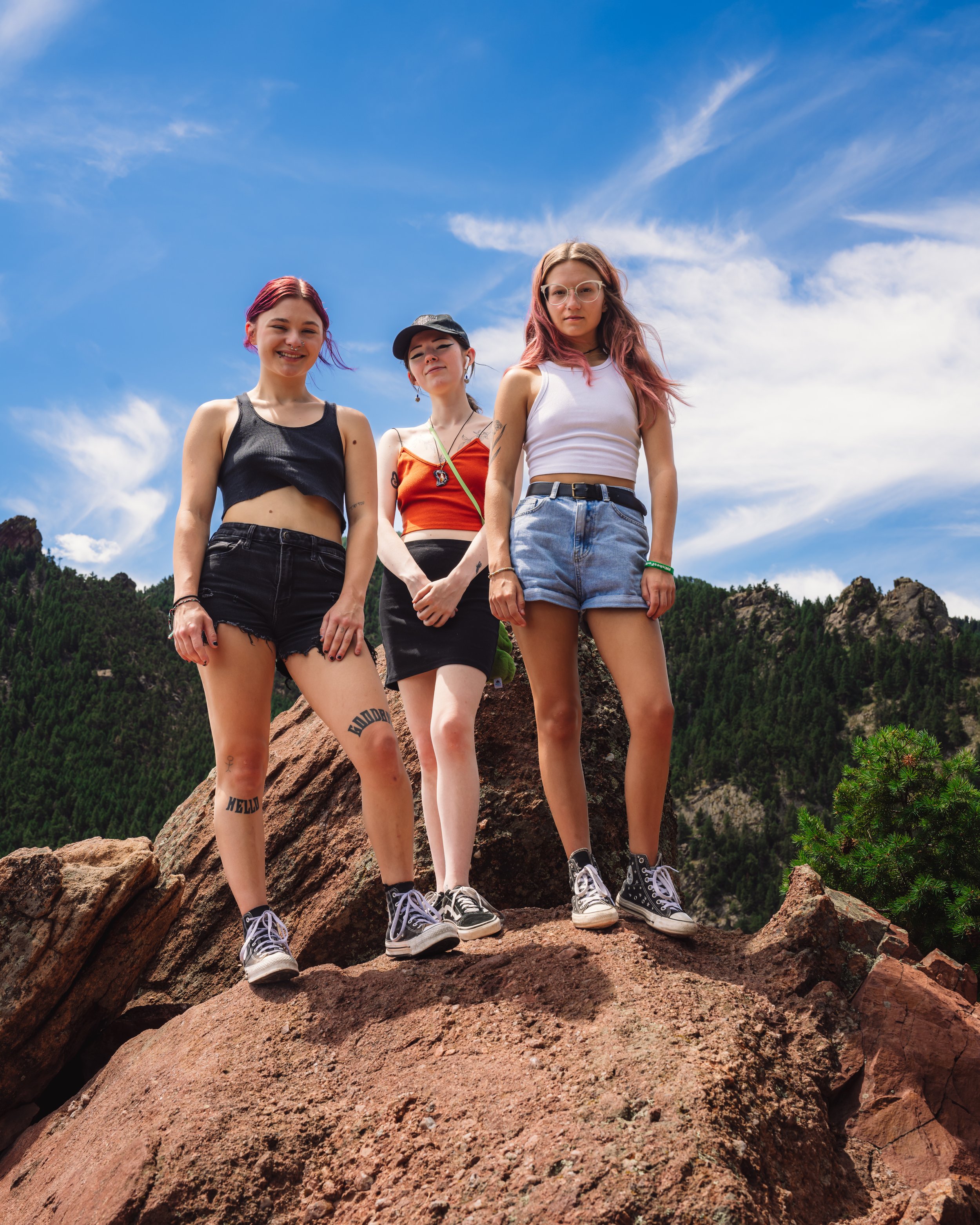 Three young women standing on rocks outdoors with mountains and trees in the background under a blue sky with clouds.