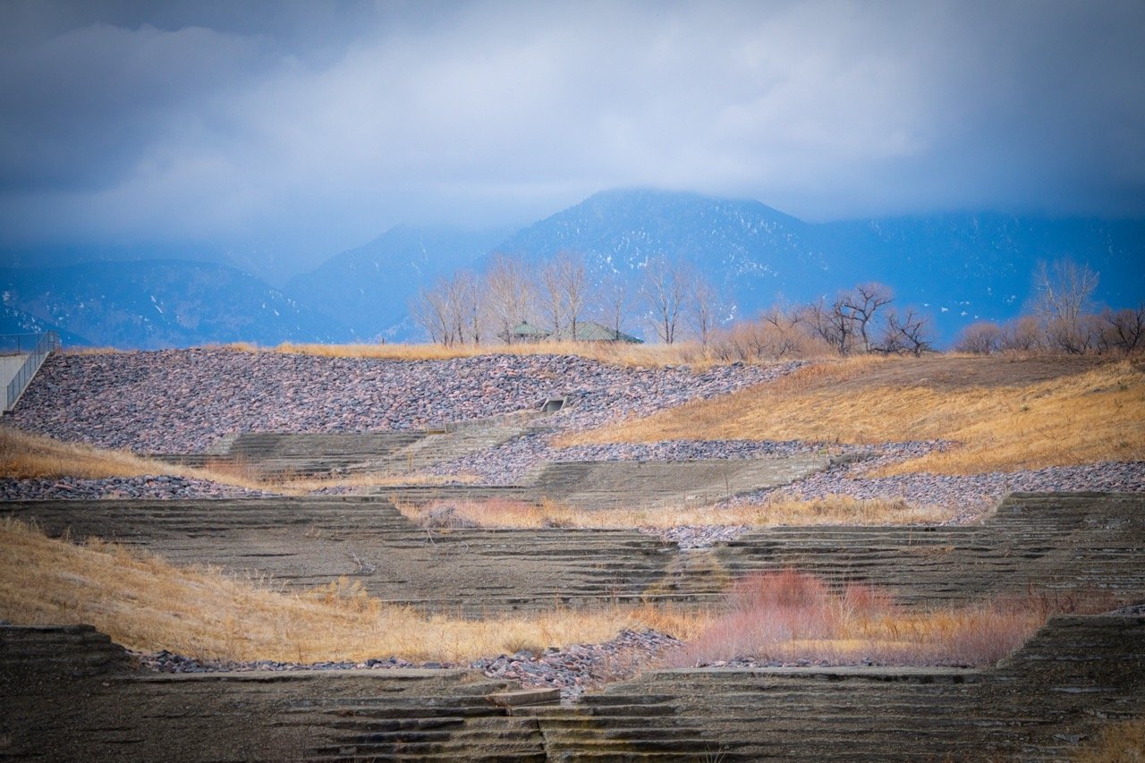 Scenic view of a terraced hillside landscape with grass, rocks, and steps leading up, with leafless trees and mountains in the background under cloudy sky in Standley Lake State Park, CO.