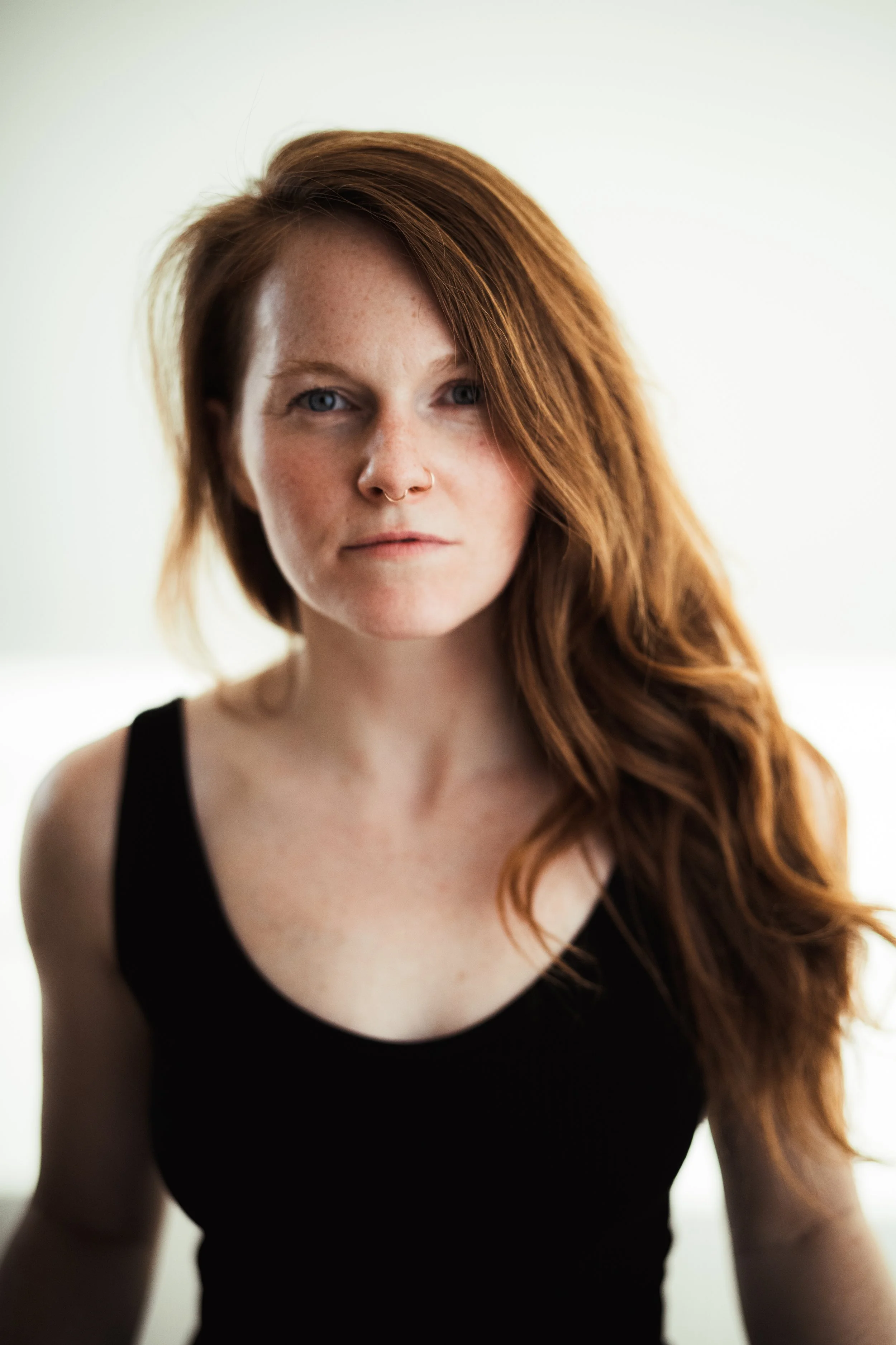 A woman with long red hair, fair skin with freckles, wearing a black tank top, looking at the camera with a serious expression.