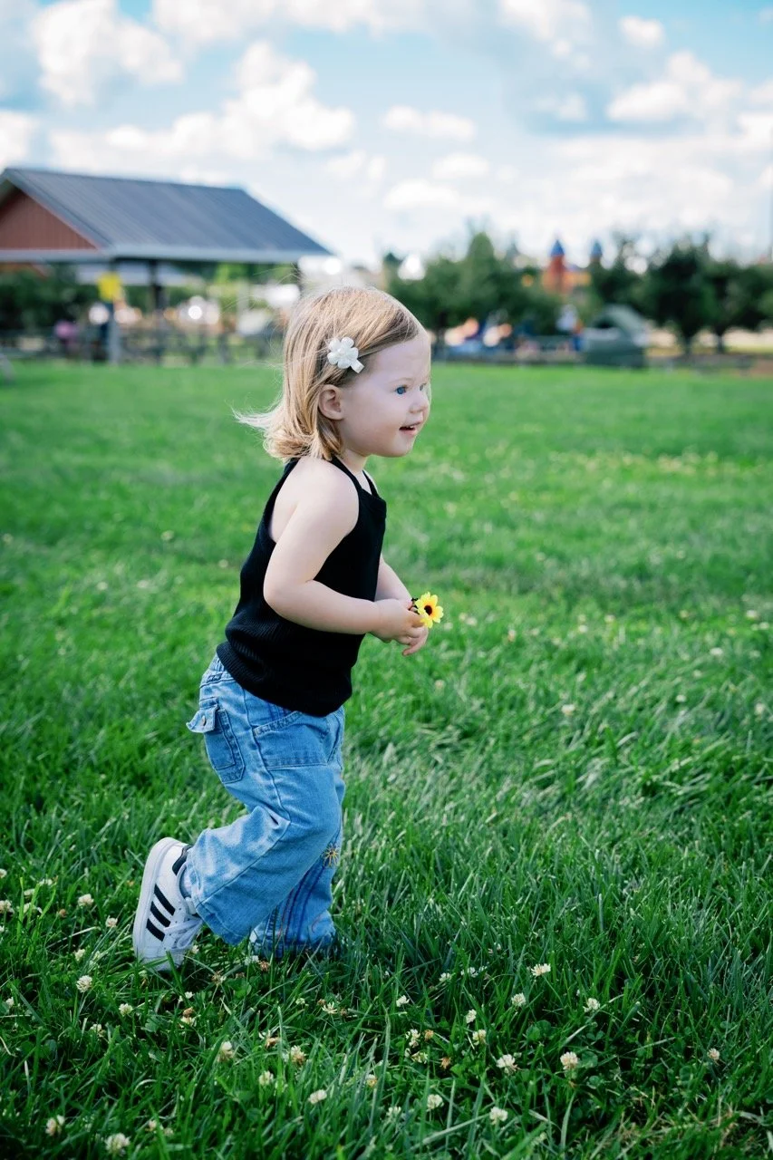A young girl with light brown hair, wearing a black tank top, blue jeans, and white sneakers with black stripes, is running on a grassy field holding a yellow flower. She has a white flower hair clip and is smiling.