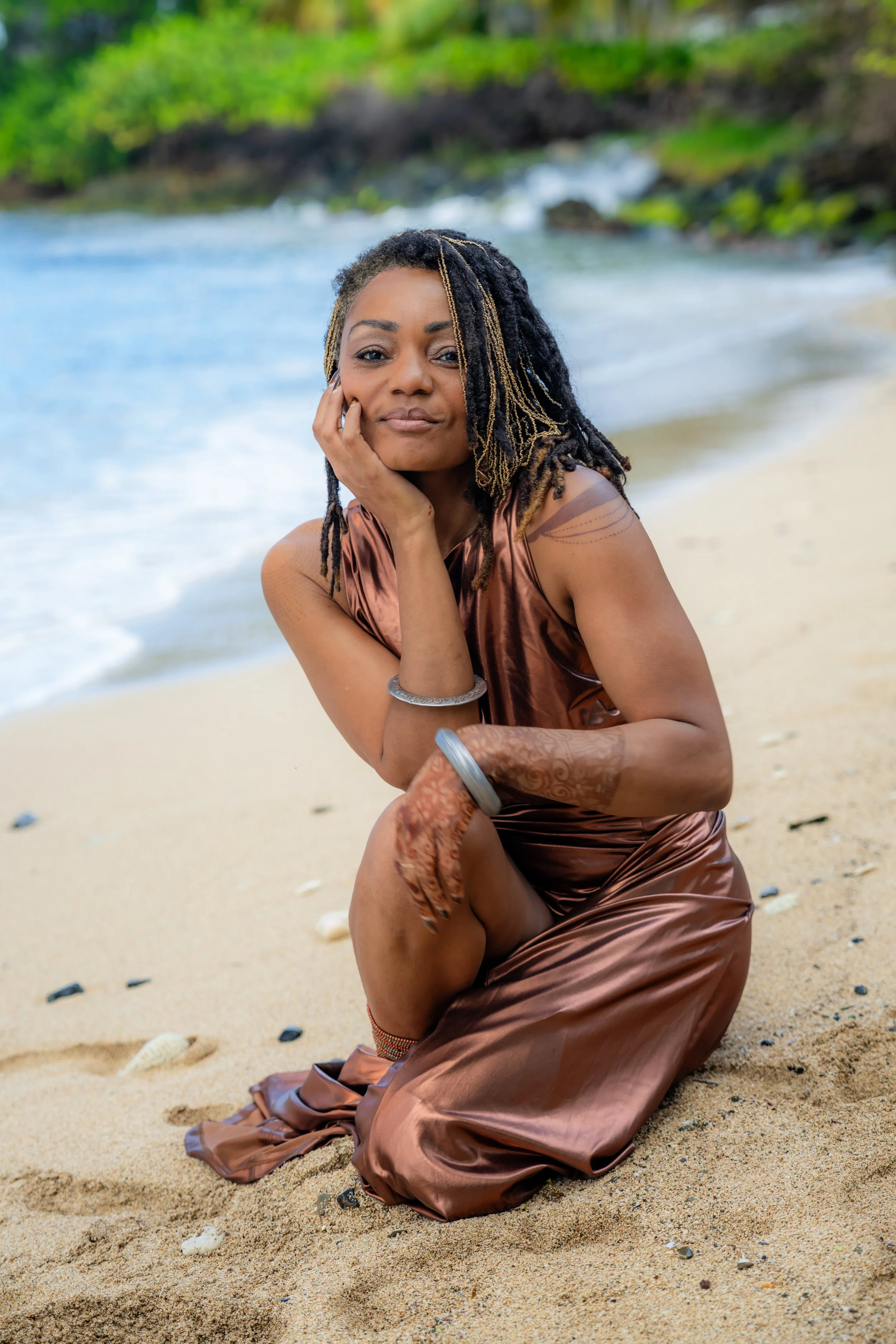 A woman with dreadlocks wearing a satin bronze-colored dress kneels on a sandy beach in St. Croix, resting her face on her hand, with the ocean in the background.