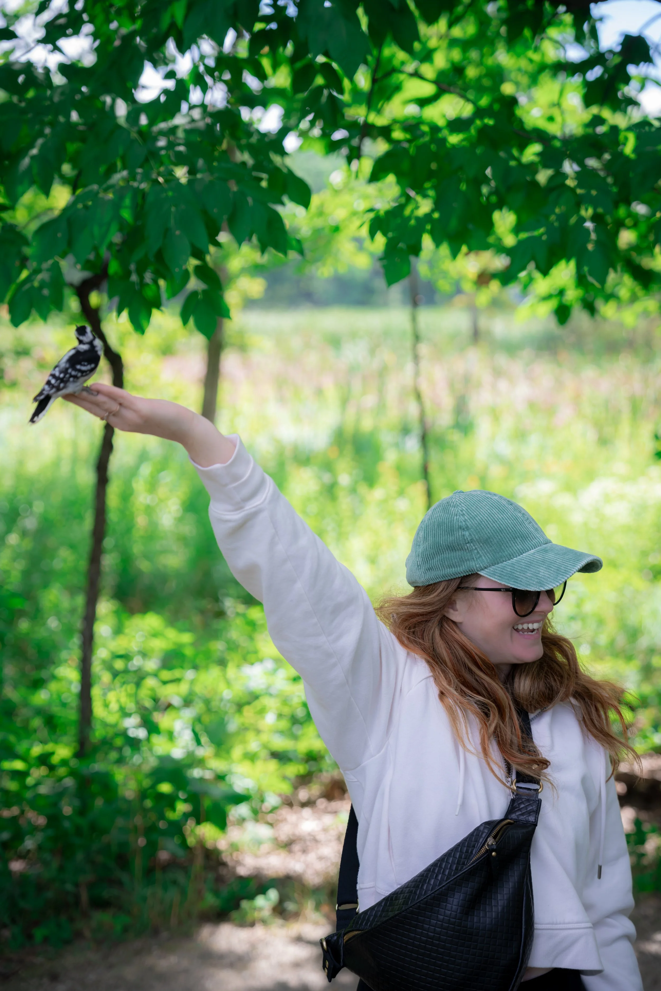Woman with glasses and a green cap smiling and reaching out her arm with a small bird perched on her hand in a lush green outdoor setting, Kensington State Park, MI.