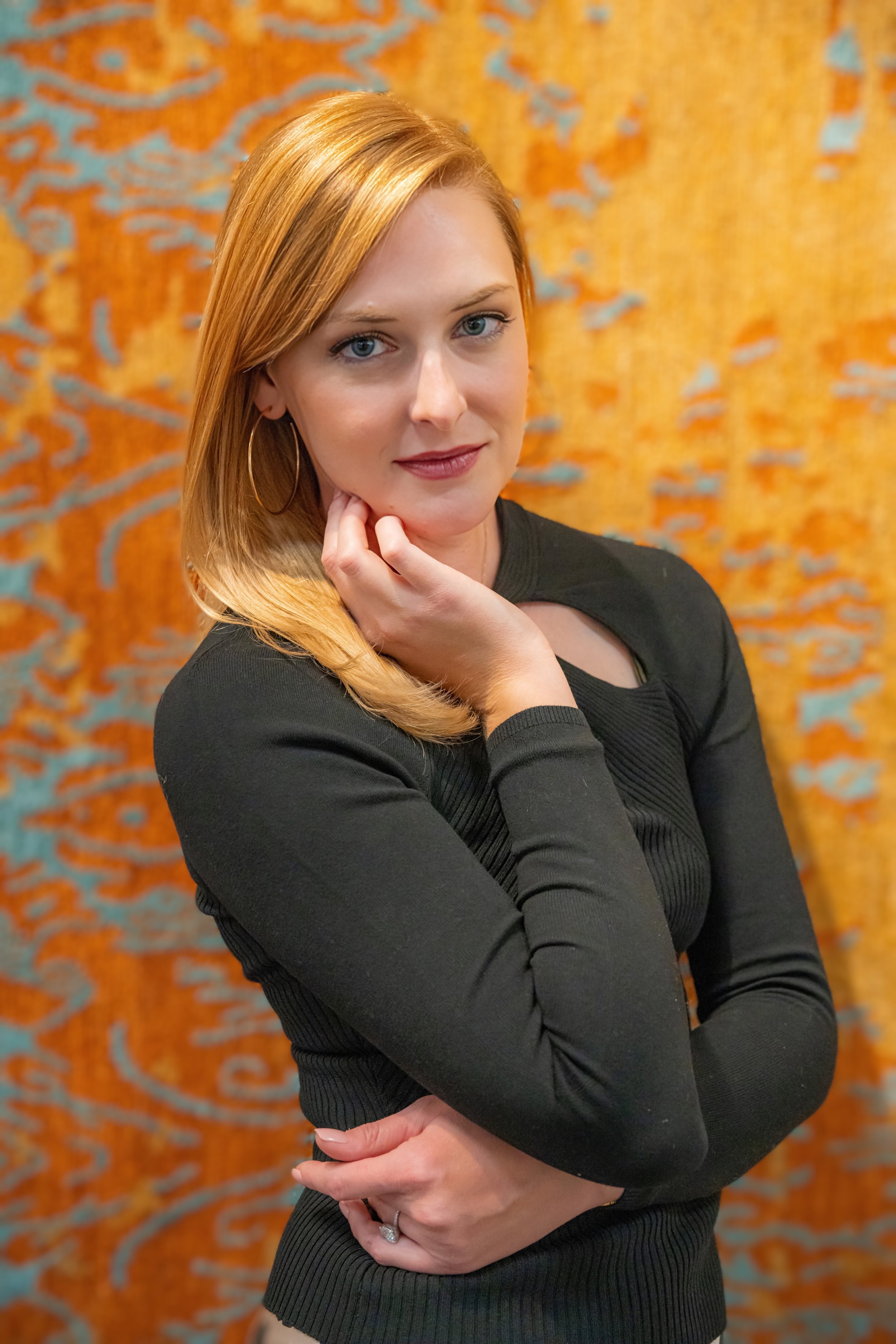 A woman with red hair and blue eyes looking at the camera, wearing a black long-sleeve top with a small cutout, standing in front of an orange and blue patterned background.