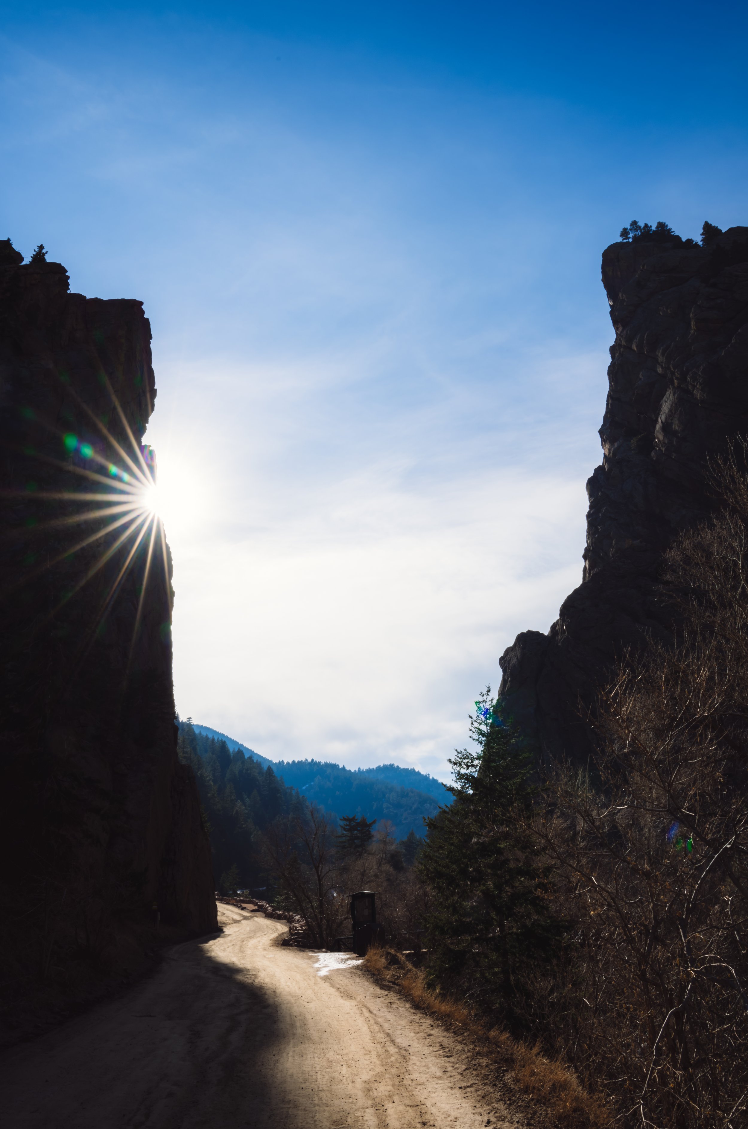 The winding road through Eldorado Canyon State Park, between tall rocky cliffs on either side, with trees and mountains in the background, and the sun shining through a gap between the cliffs.