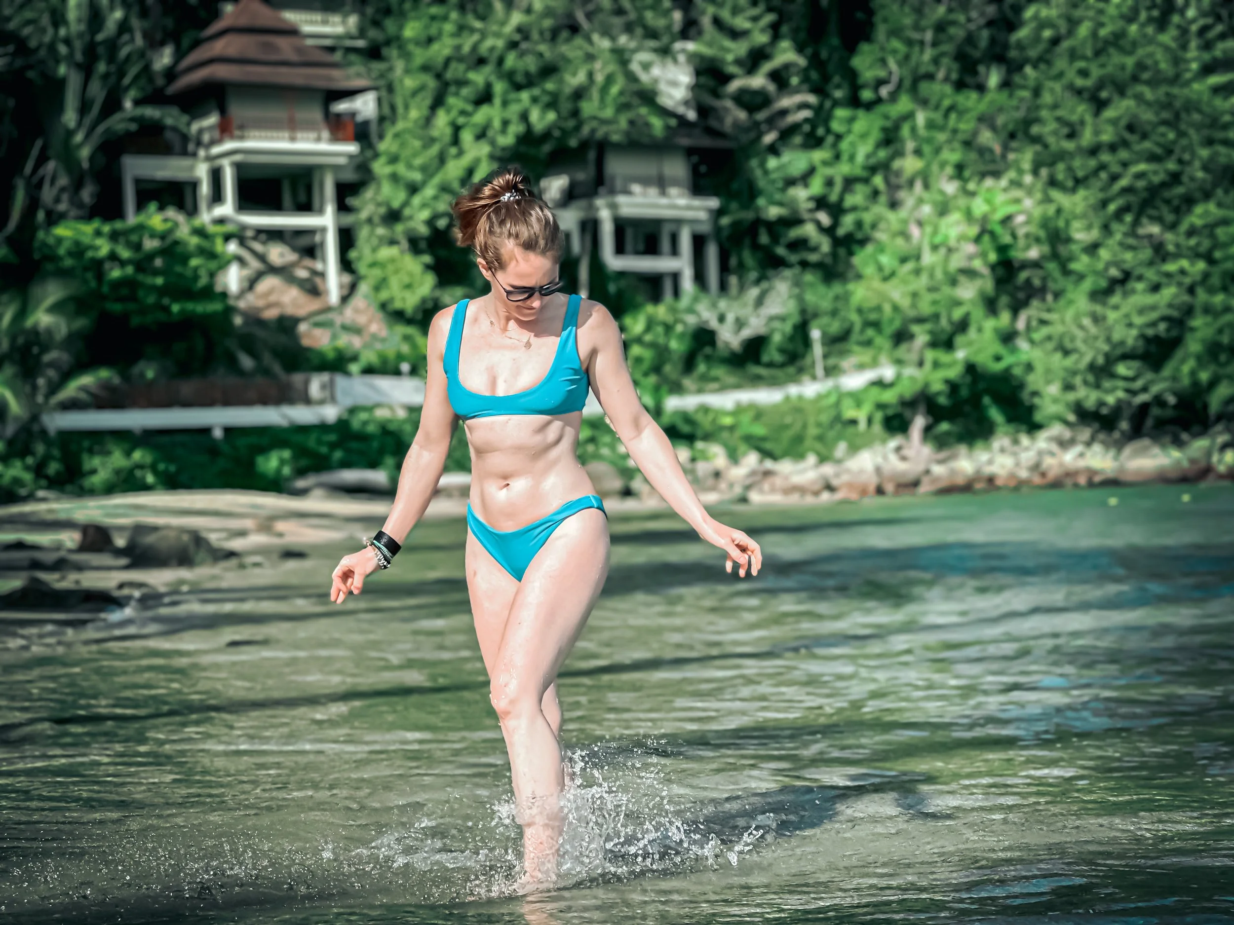 Woman in a blue bikini walking in shallow water near a rocky shoreline in Phuket, Thailand with green trees and small wooden houses in the background.