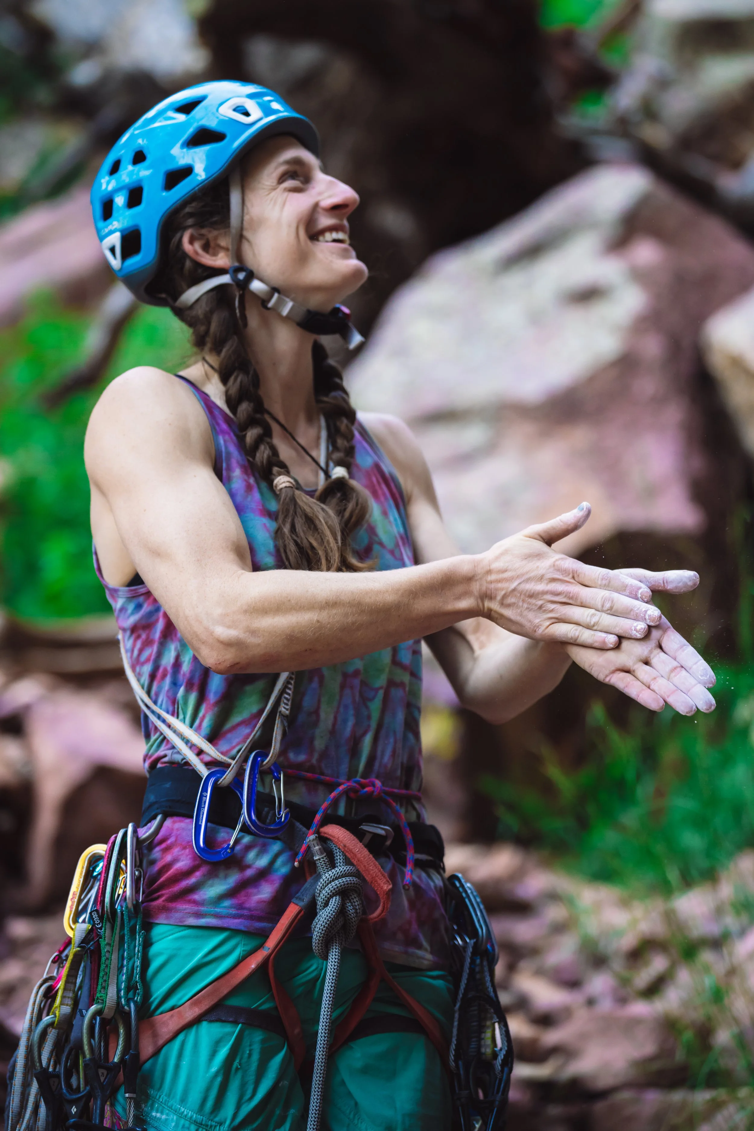 Becca Droz, wearing climbing gear, a blue helmet, and a sleeveless shirt, smiling as she prepares to begin. 