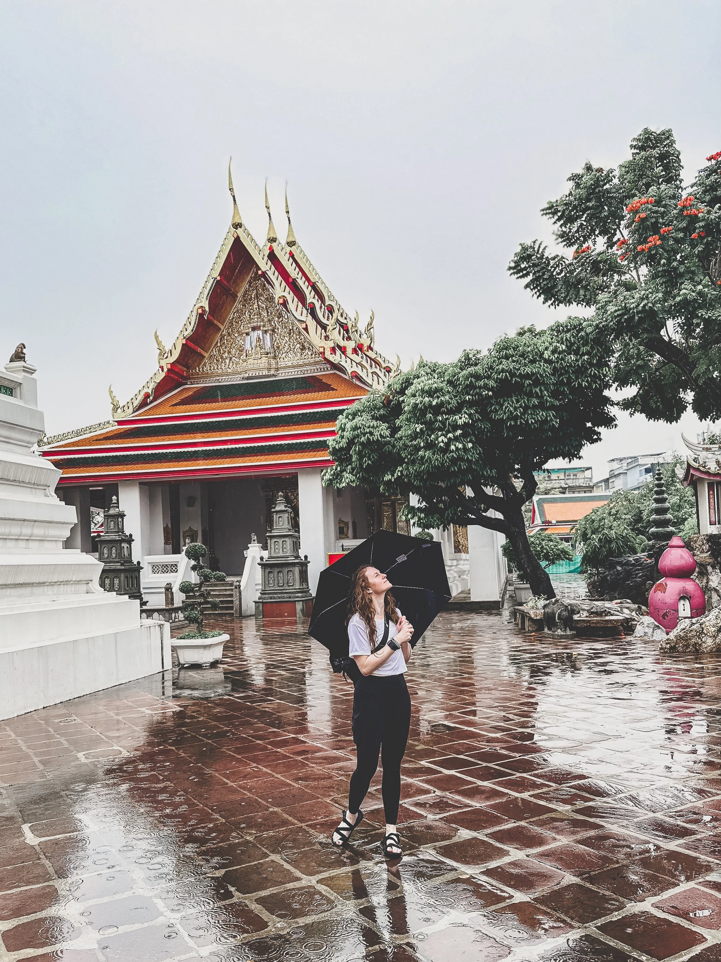 A woman holding a black umbrella stands on a rainy, reflective stone ground in front of a traditional building of the Wat Pho temple with ornate roof Design and decorative elements, surrounded by trees and greenery.