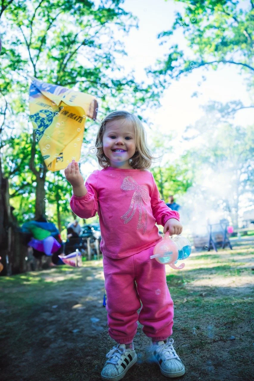 A young girl with blonde hair smiling at the camera, wearing a pink outfit with a sparkly figure, holding a bubble blower, outdoors in a park with trees and blurred people in the background.