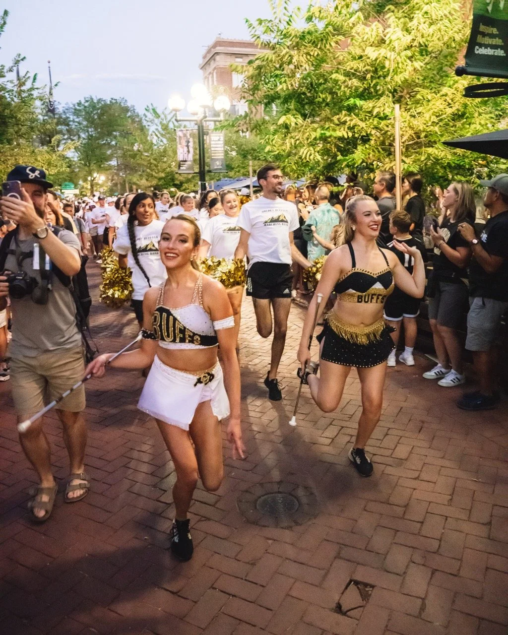 Cheerleaders from the University of Colorado in black and white uniforms with gold accents performing in a parade on a Boulder, CO street, spectators lining the sidewalk.