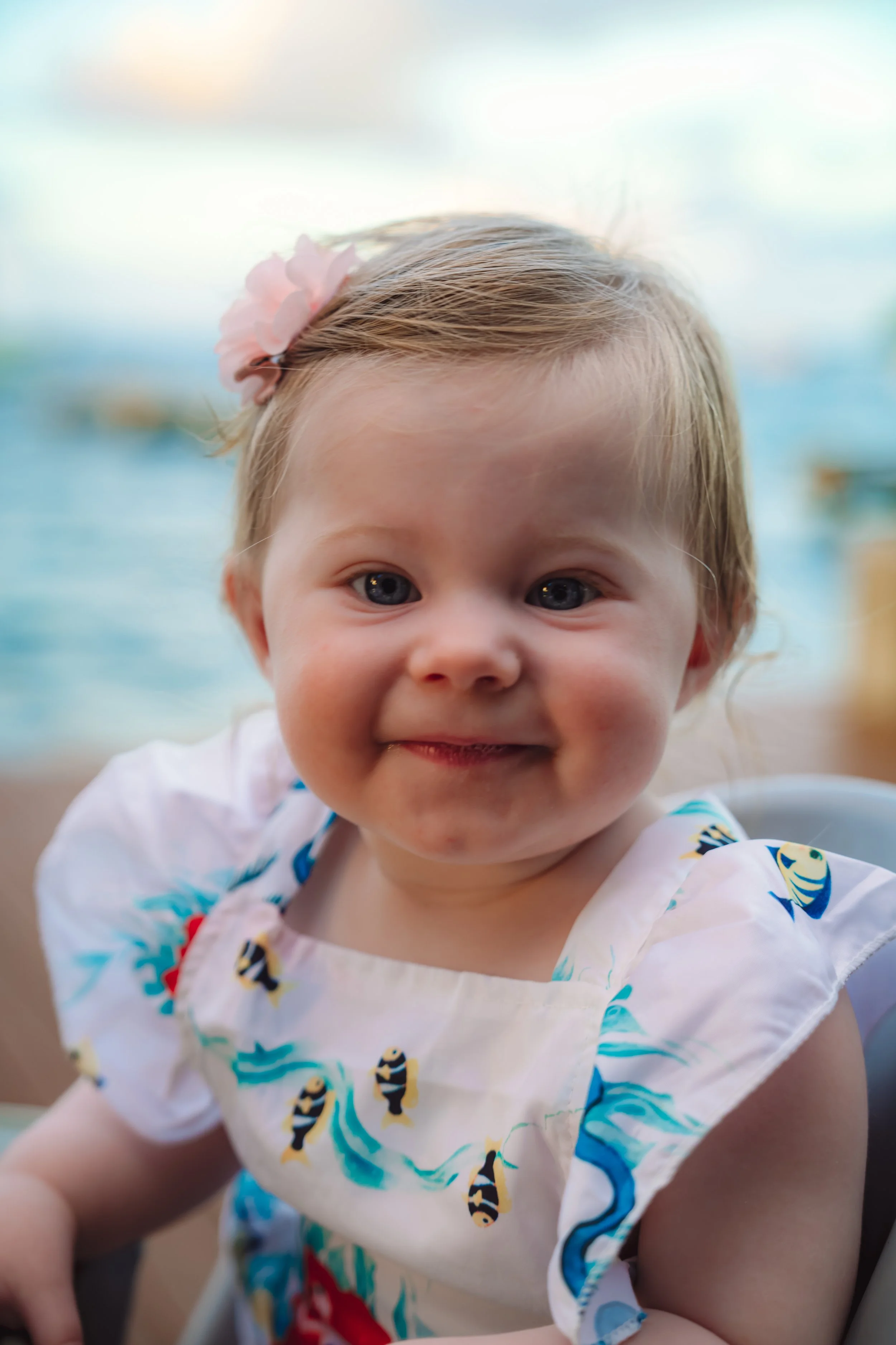 Close-up of a smiling young girl with light brown hair and a pink flower hair clip, sitting outdoors near a body of water, wearing a white dress with colorful fish print.