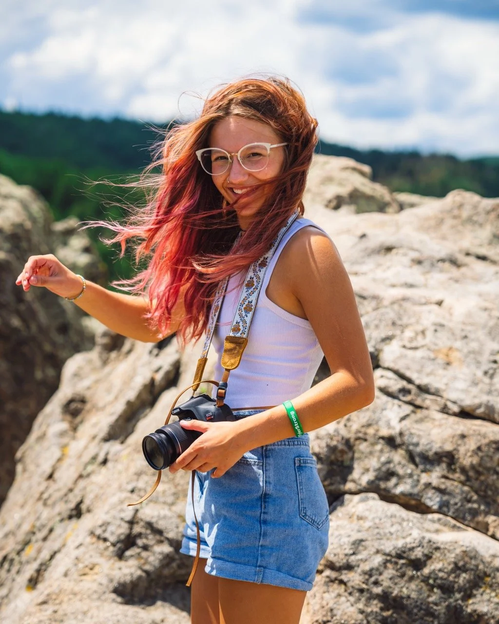 A young woman with pink hair and glasses smiling outdoors on Flagstaff Mountain in Boulder, holding a camera.