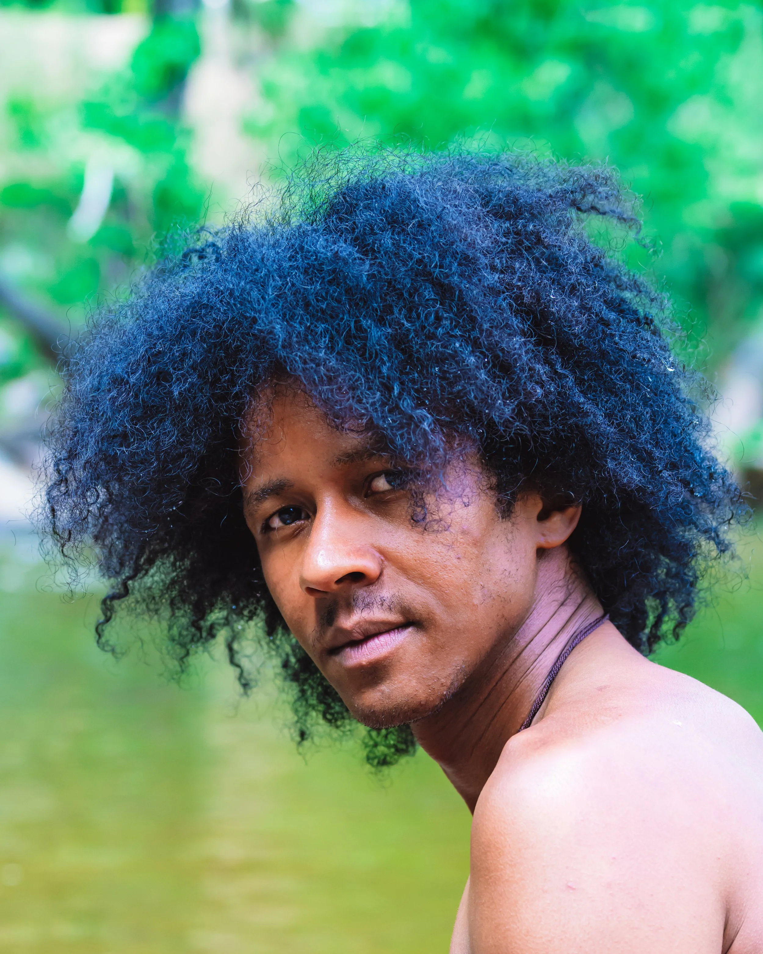 A young man with voluminous curly black hair and tan skin, shirtless, sitting near Boulder Creek with green blurred background.