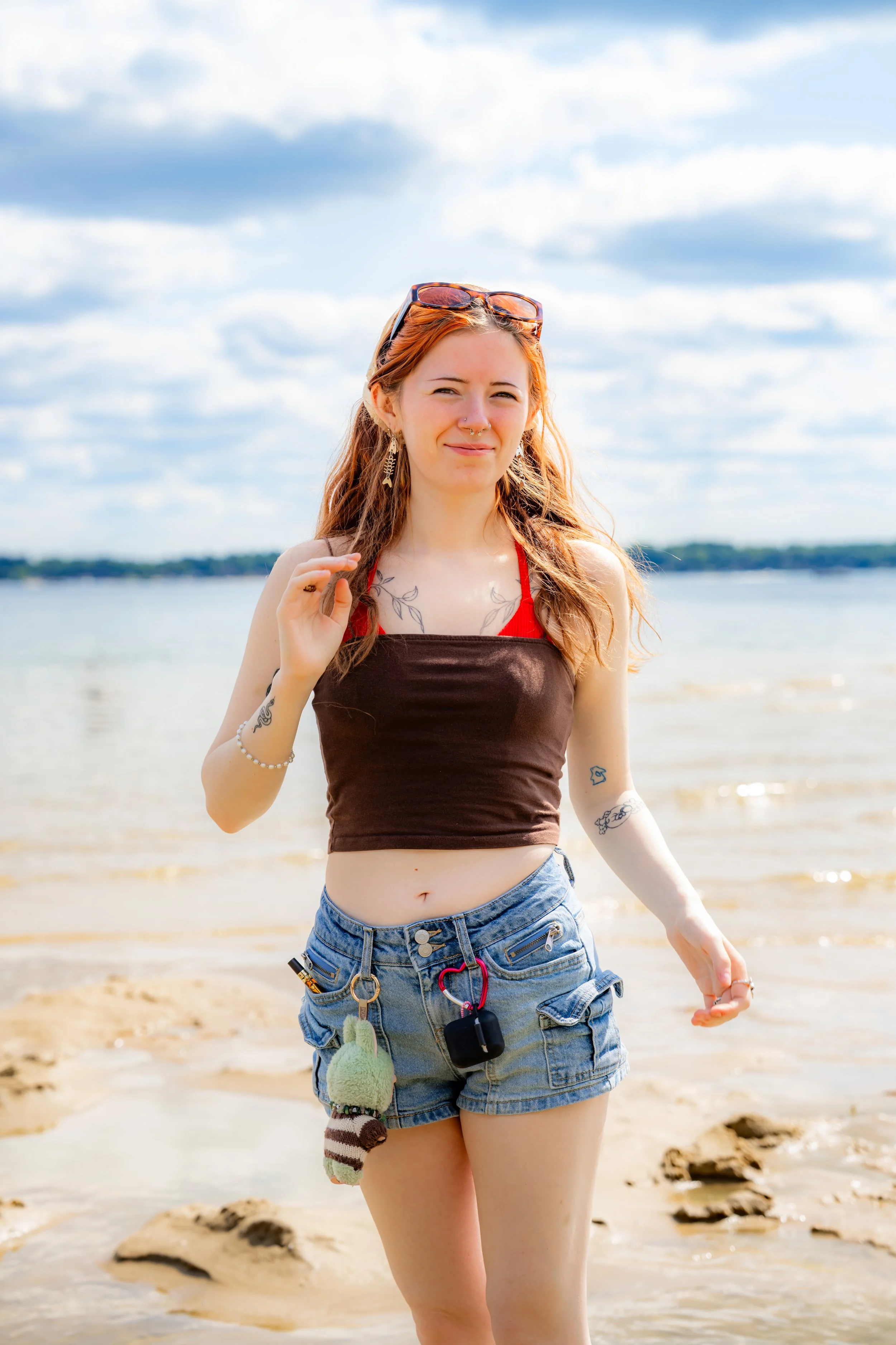 A young woman with wavy reddish-brown hair, sunglasses on her head, and tattoos on her arms and chest, standing on a sandy beach with water and a cloudy sky in the background, wearing a brown tank top with red straps and denim shorts.
