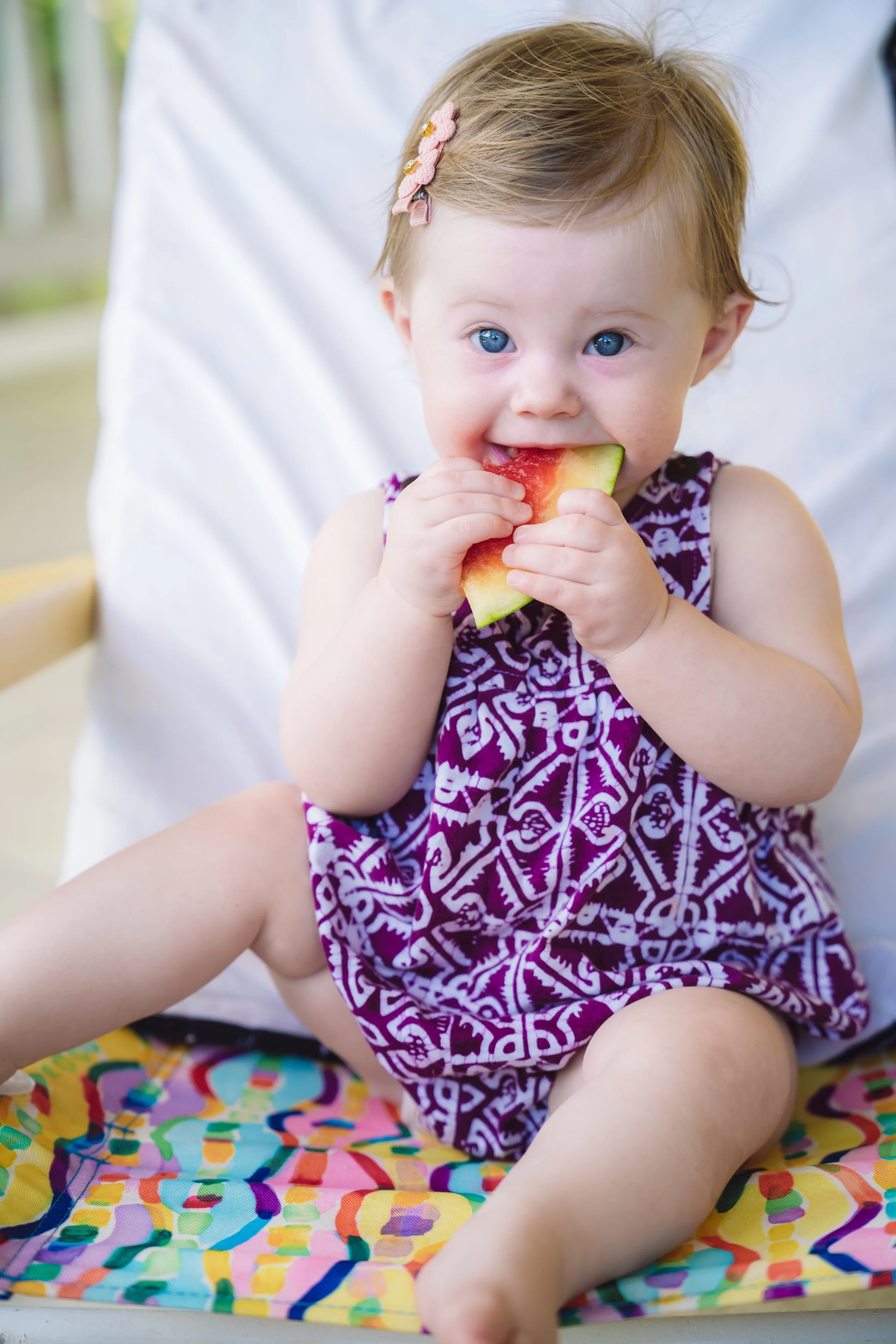 A young girl with blue eyes and a pink flower hair clip is sitting on a colorful blanket, eating a slice of watermelon in St. Croix.