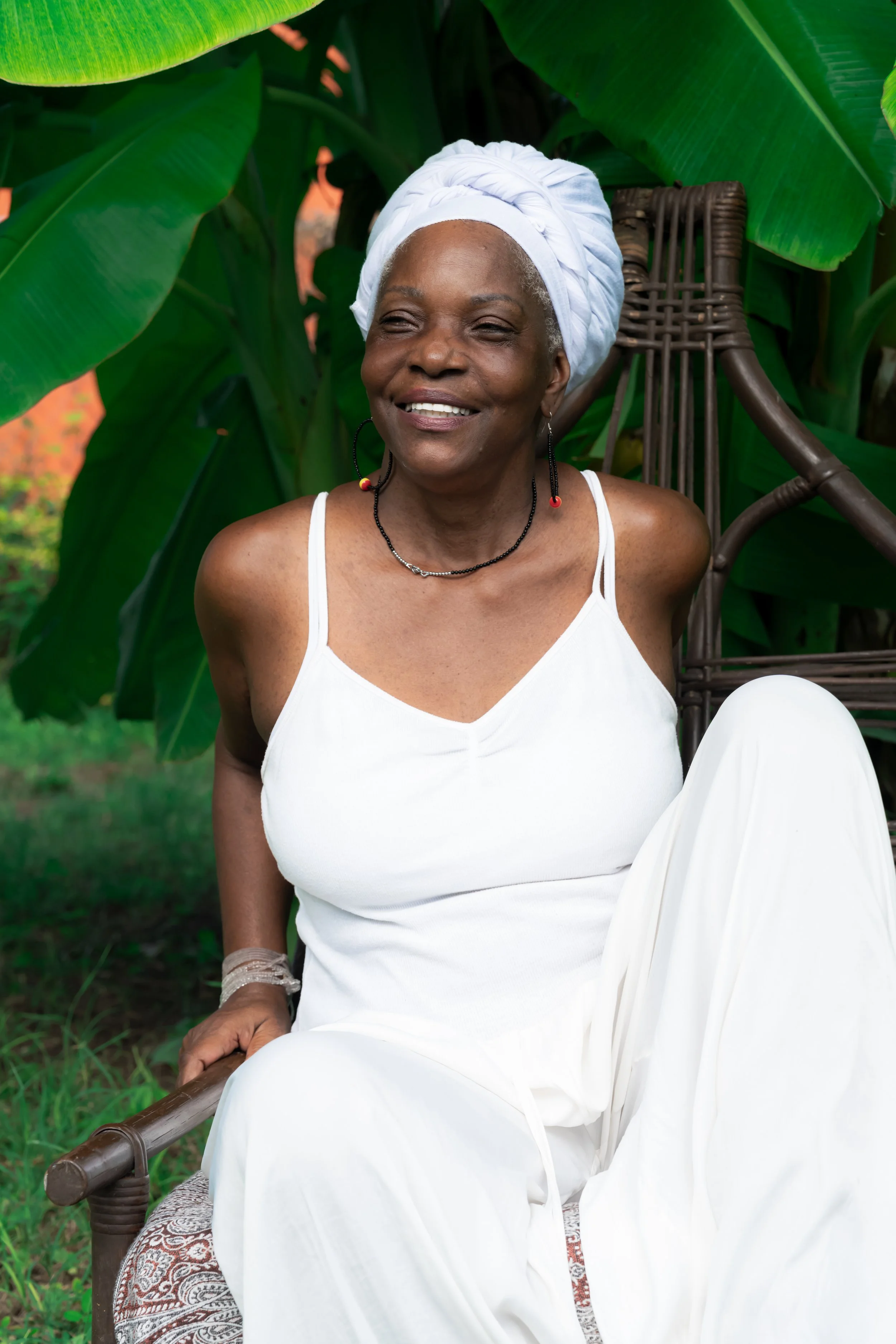 A smiling older woman with a white headwrap, wearing a white sleeveless dress, sitting on a wooden chair outdoors amidst lush green banana leaves.