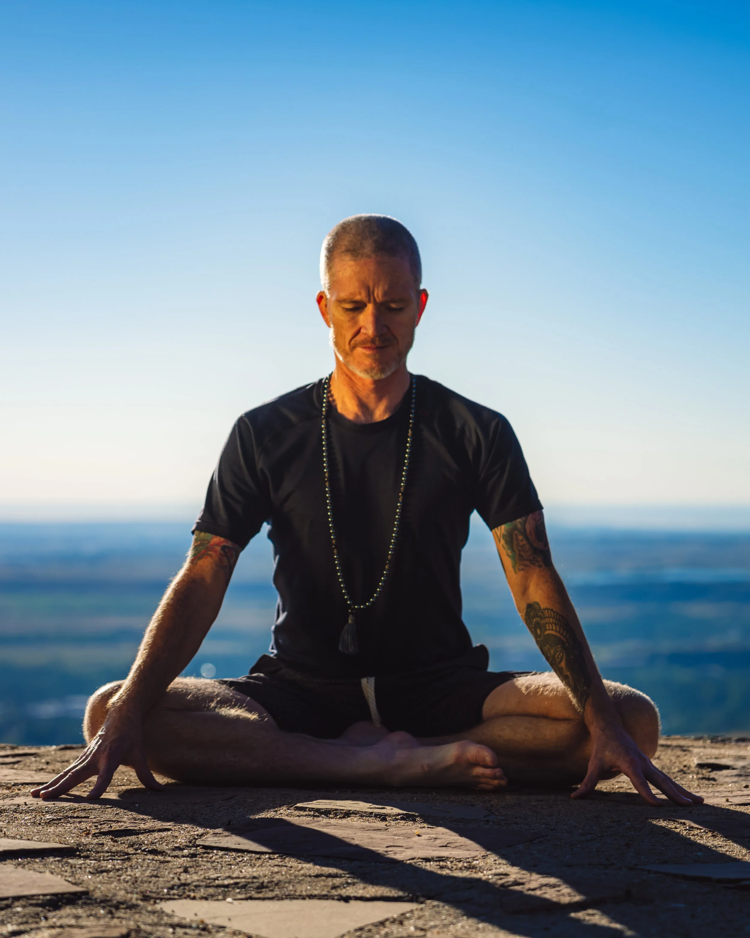Rob Loud practicing yoga on the stone surface of Flagstaff Mountain, sitting cross-legged with hands resting on the knees, eyes closed, under a clear blue sky overlooking the city of Boulder, CO.