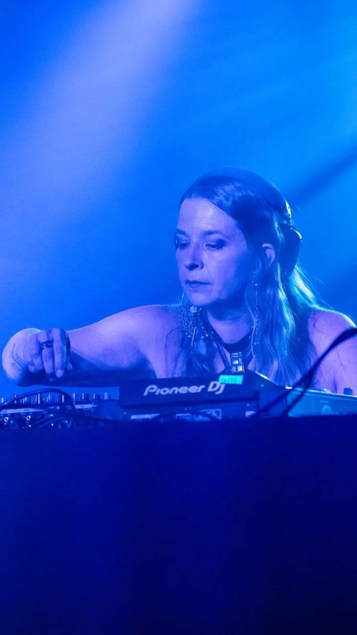 Magnetik Moon with long wavy hair, wearing a sleeveless top, operating a Pioneer DJ equipment in a dimly lit, blue-toned Cervantes' Other Side.