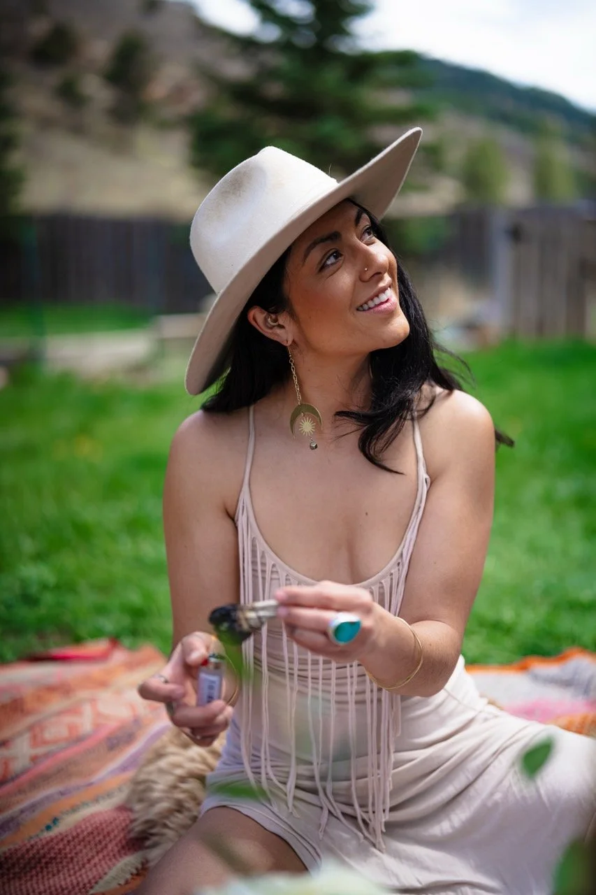A woman sitting on a blanket outdoors, holding a lighter and sage, wearing a wide-brimmed hat, a sleeveless top with fringe, and smiling while looking upward. The background shows trees, grass, and a wooden fence.