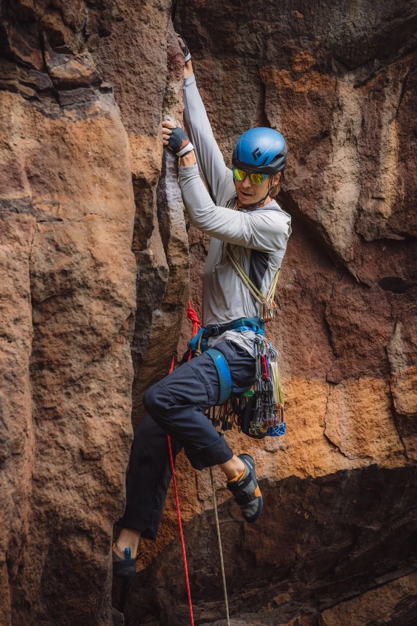 A male rock climber wearing a blue helmet, sunglasses, and climbing gear is ascending a narrow, rocky canyon wall.
