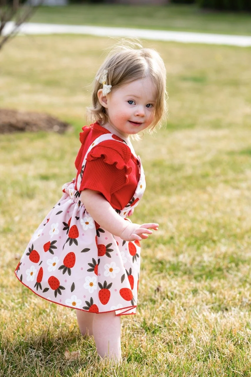 A young girl with blonde hair, wearing a red shirt with ruffled sleeves and a strawberry-printed dress, is standing on grass outside.