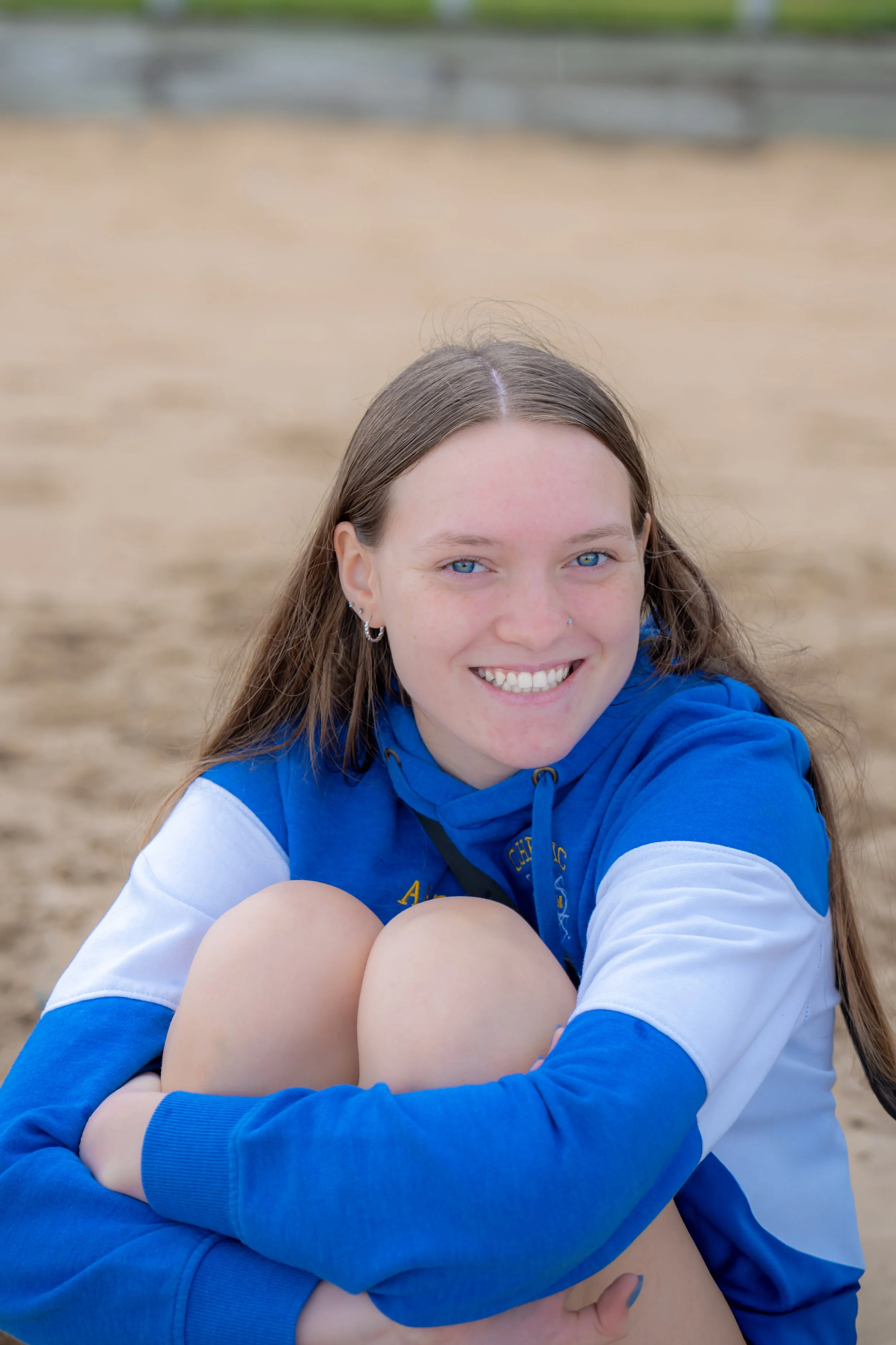 A young woman with long brown hair and blue eyes sitting on a sandy surface, smiling at the camera, wearing a blue and white hoodie.