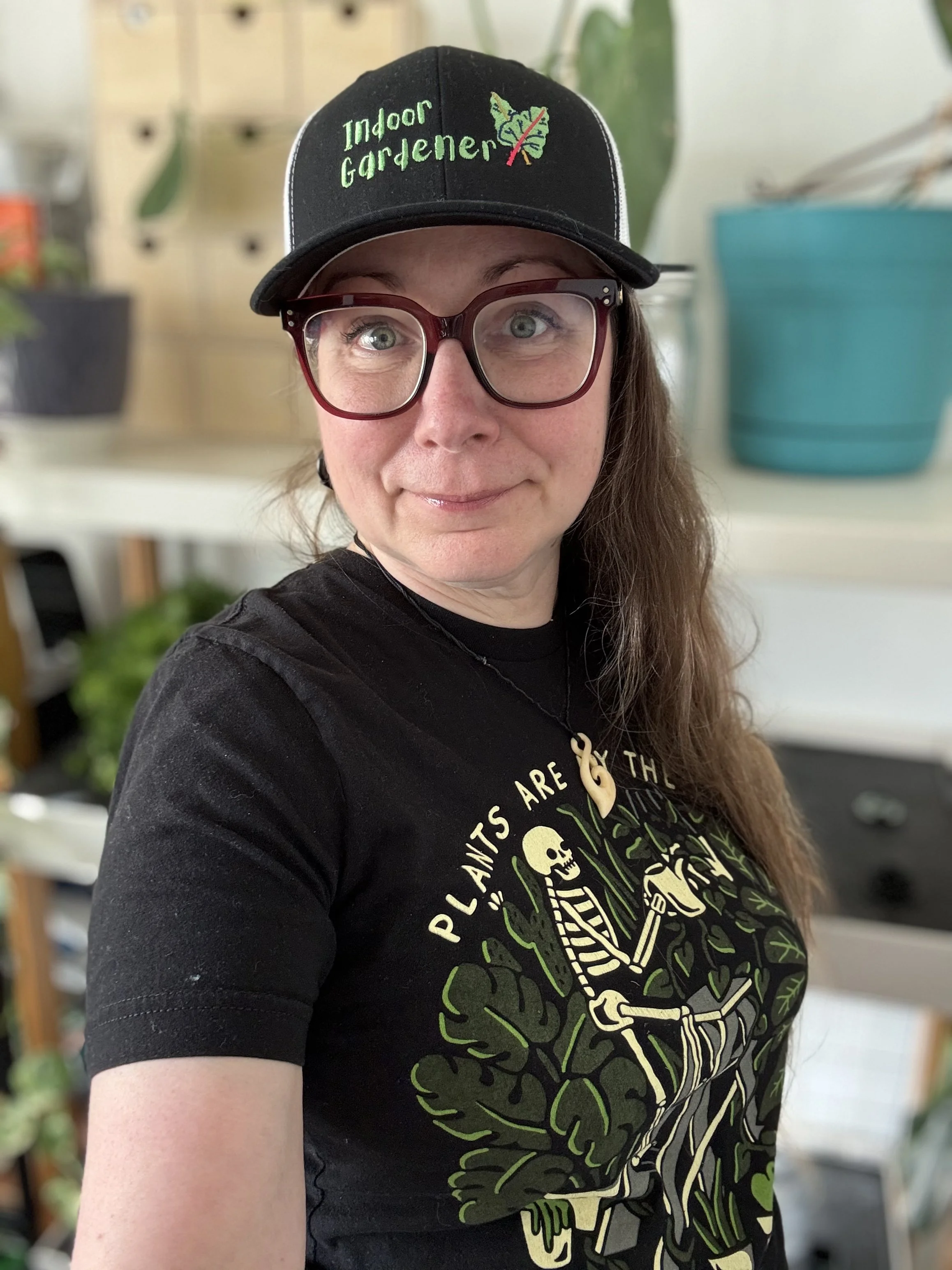 A woman with green eyes and glasses wearing a black baseball cap with green embroidered text that says 'Indoor Gardener' and a graphic of a leaf crossed out; she is also wearing a black T-shirt with a skeleton and green plants design, standing in front of a background with plants and shelves.