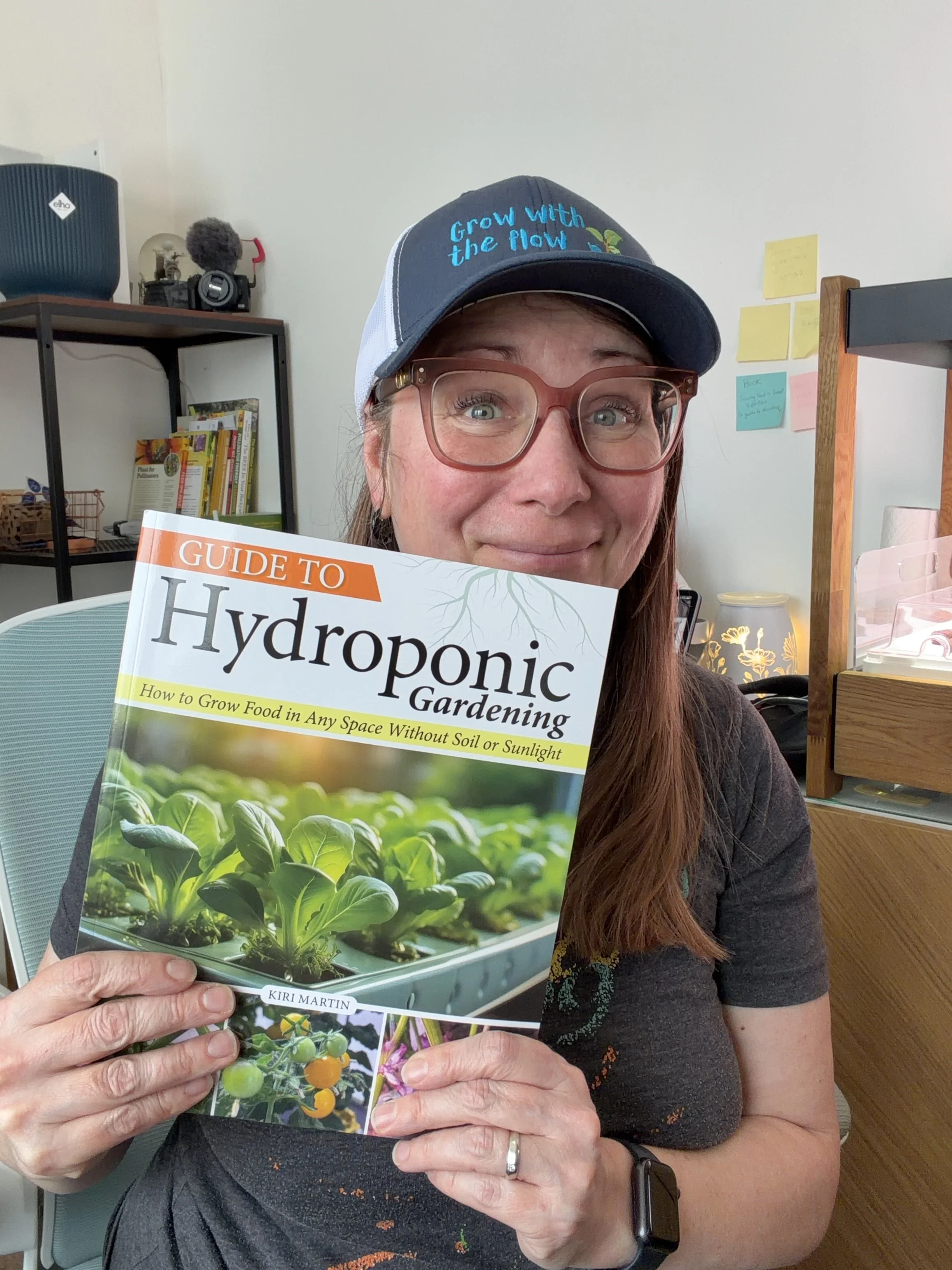 A woman wearing glasses and a cap that says 'Grow with the flow.' She is holding a book titled 'Guide to Hydroponic Gardening' by Kiri Martin. The background shows a room with shelves, books, and notes on the wall.