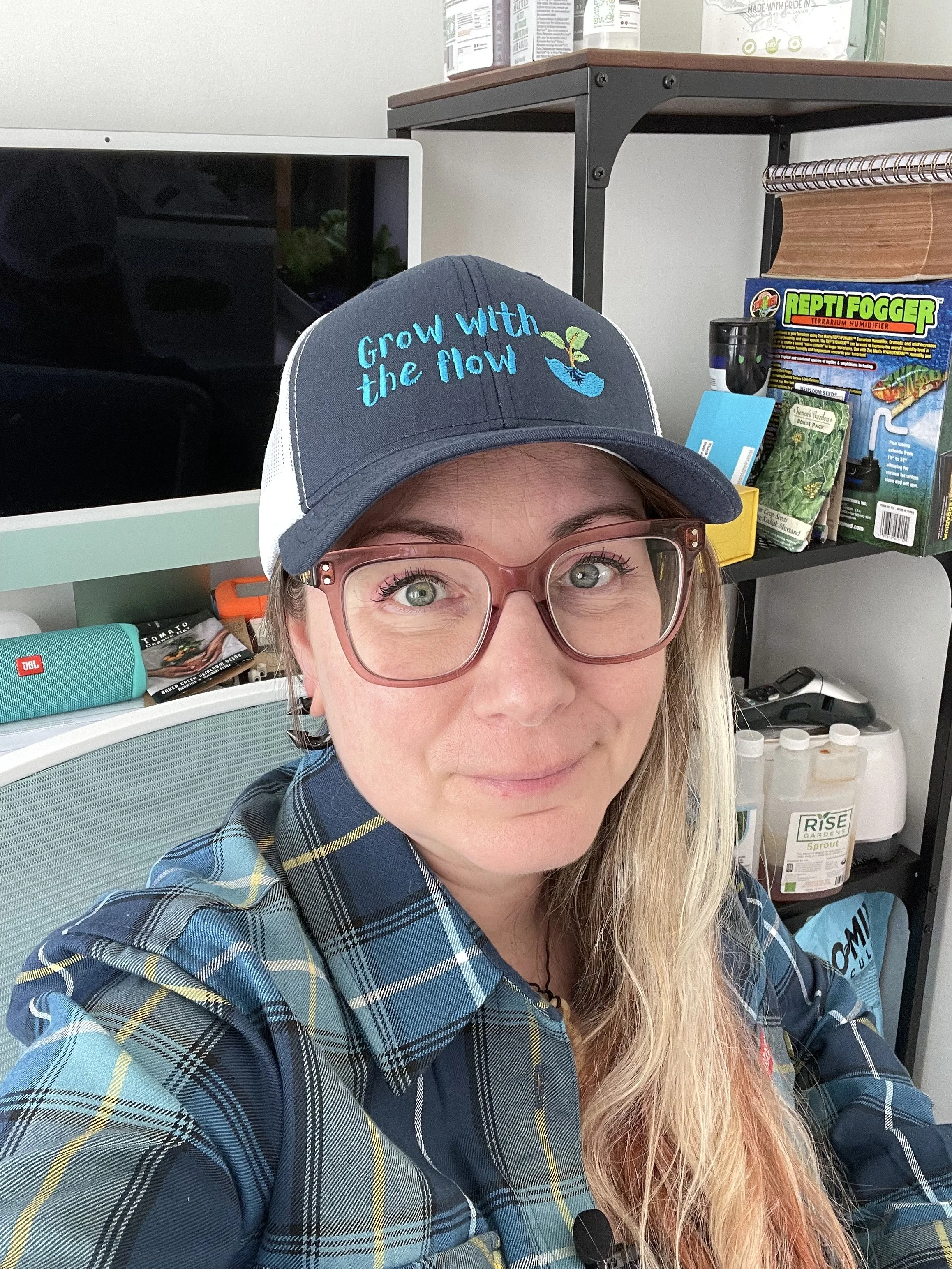 Close-up of a woman with blonde hair wearing glasses and a baseball cap that says "Grow with the flow." She is wearing a plaid shirt and sitting in an office or lab environment with shelves containing various supplies and a black computer monitor behind her.