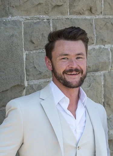 A man with dark hair and a beard, wearing a white suit and shirt, standing in front of a stone wall, smiling.