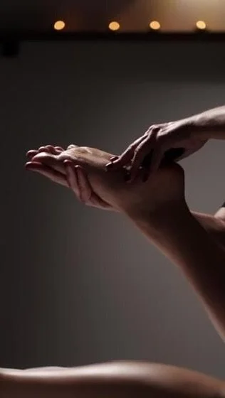 A person receiving a massage on their hand or wrist with their palm facing up, in a dimly lit room with small overhead lights.