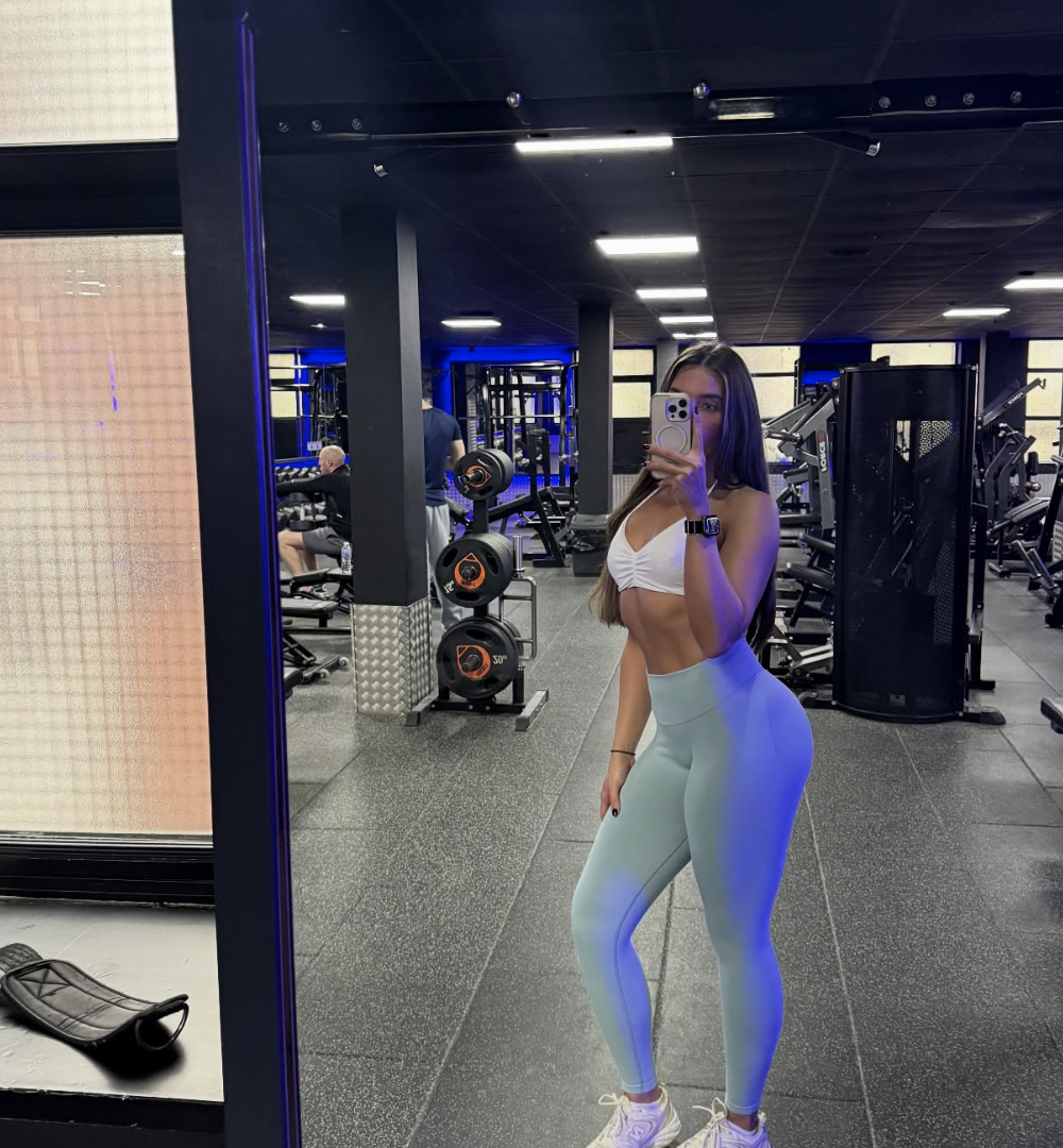 Close-up photo of a young woman with dark hair styled in a messy bun, smiling, wearing a white top, and standing against a plain white background.