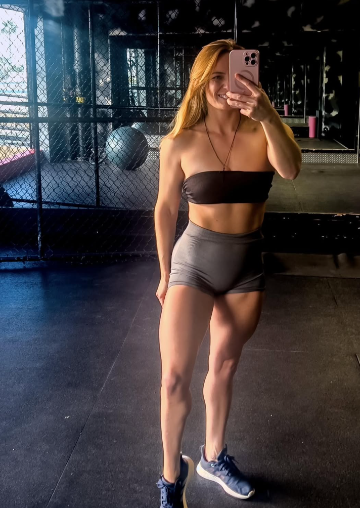 Young woman standing outdoors in a park with green grass, trees, and a hillside in the background, wearing a plaid blazer and a brown top.