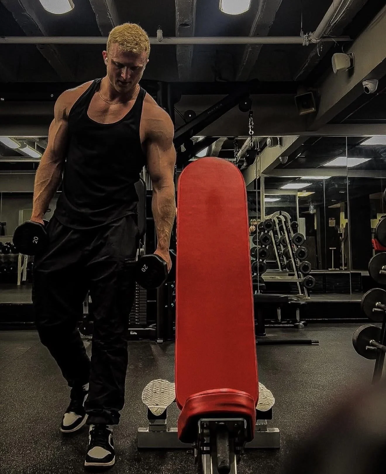 A young man with dyed blonde hair is working out in a gym, lifting dumbbells, surrounded by various gym equipment and mirrors.