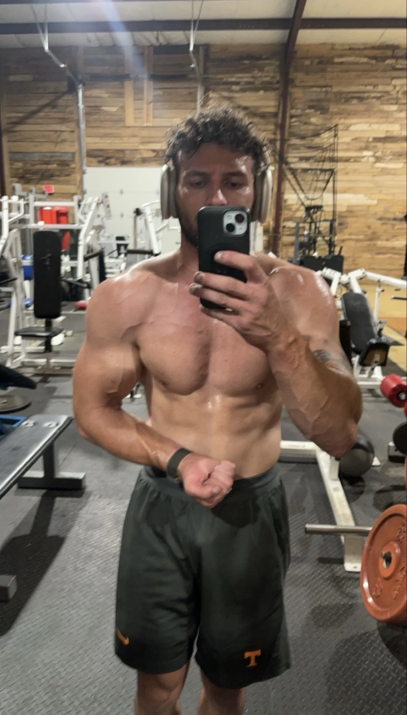 A man taking a shirtless selfie in a gym mirror, flexing his left arm, wearing headphones, black shorts with an orange logo, with gym equipment in the background.