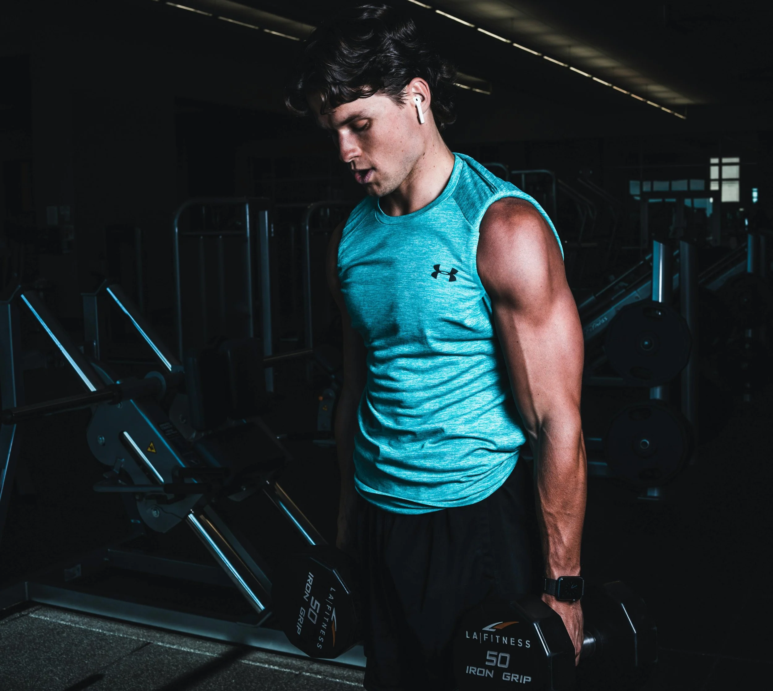 A young man with dark curly hair wearing a light blue Under Armour athletic shirt, black shorts, earphones, and a smartwatch, performing a bicep curl with 50-pound dumbbells in a dimly lit gym.