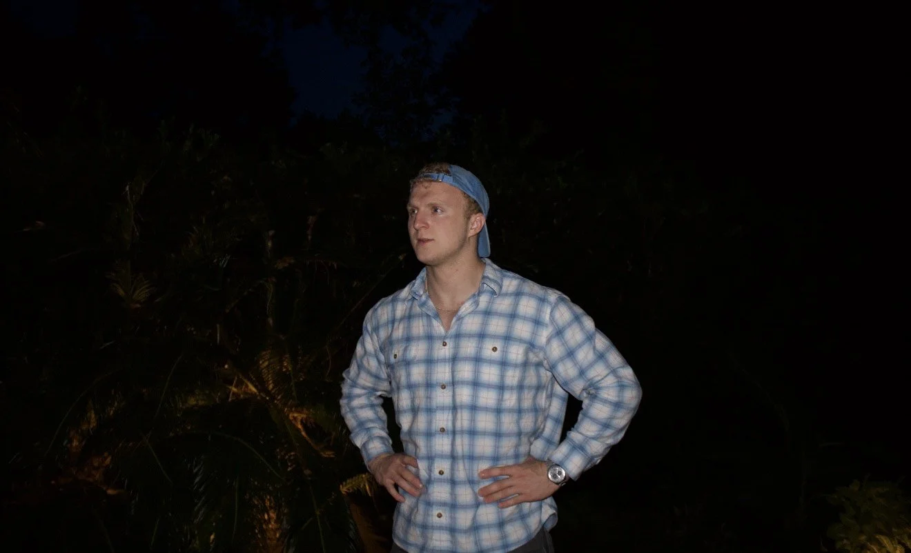 Young man wearing a blue plaid shirt and backward baseball cap standing outdoors at night with hands on hips.