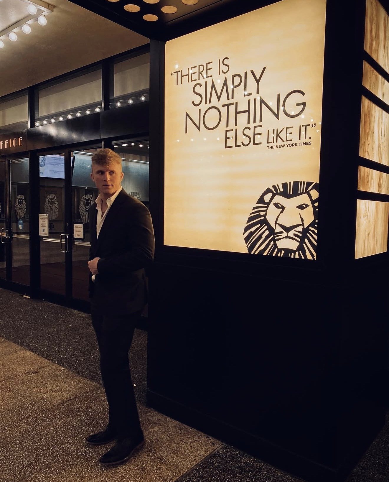 A man in a black suit standing outside a building, near a large illuminated sign with a quote and a lion logo. The quote reads, 'There is simply nothing else like it.'