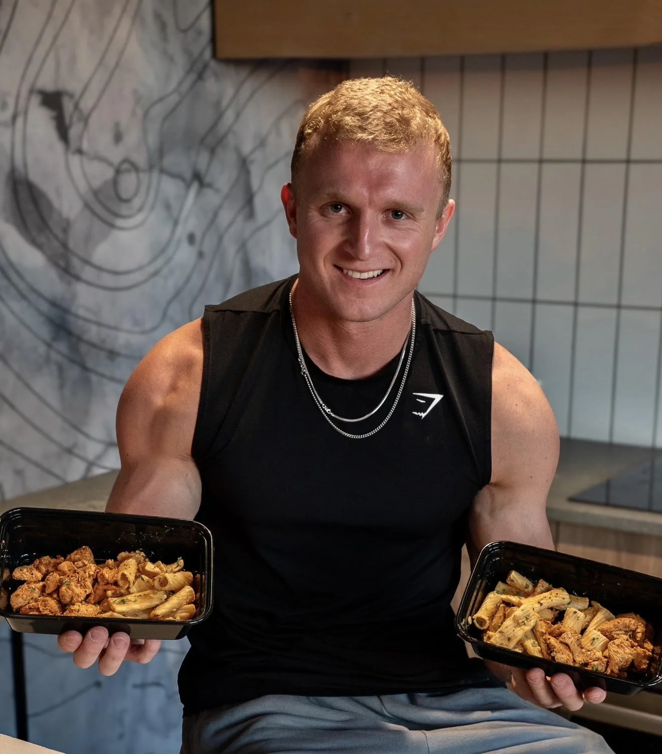 A smiling young man with short blond hair, wearing a black sleeveless athletic shirt and silver necklaces, holding two black food containers filled with pasta and meat, sitting in a modern restaurant with a gray tiled wall and abstract artwork in the background.
