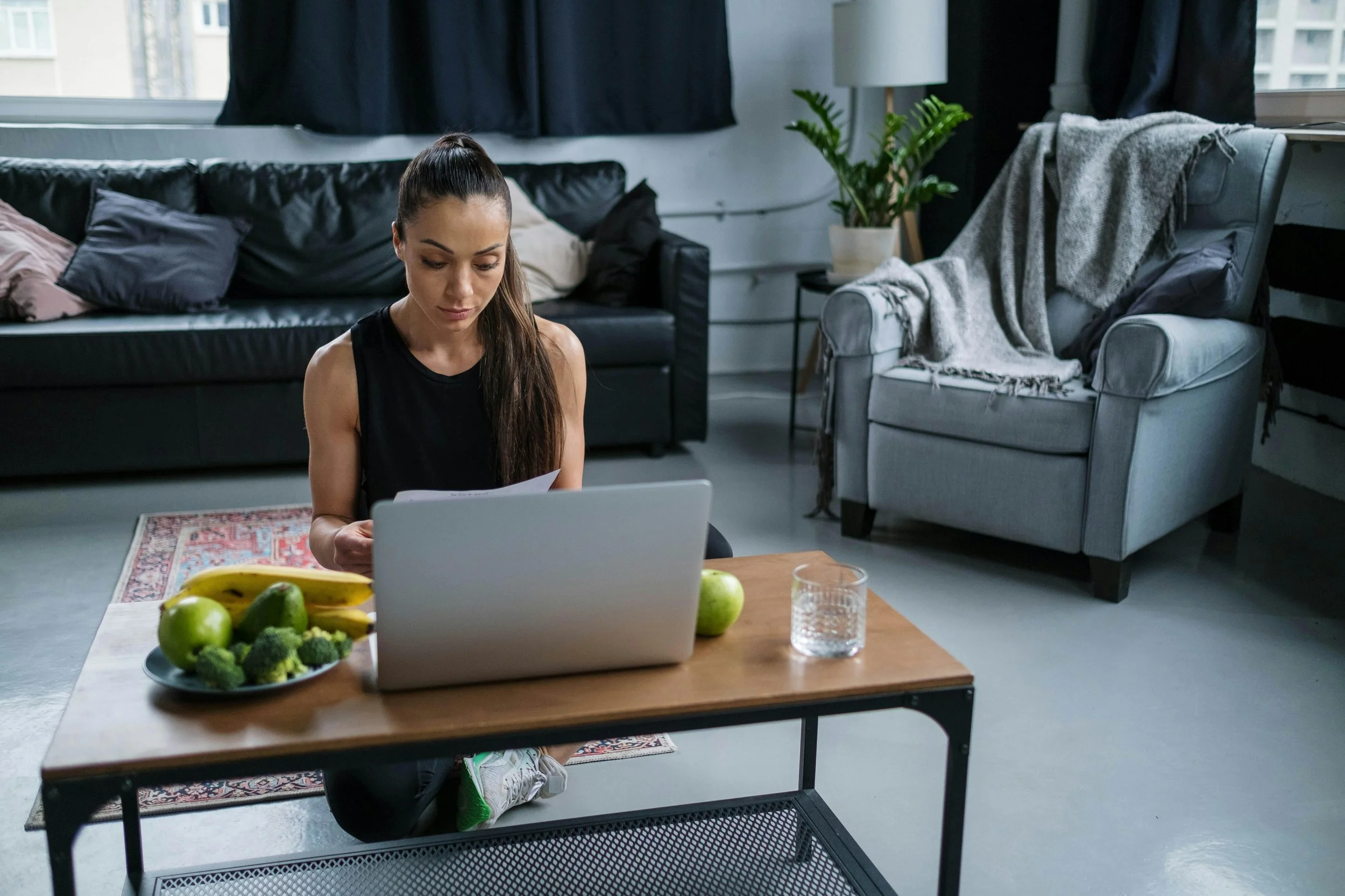 Woman sitting on the floor in front of a laptop, with a plate of bananas, apples, and broccoli, in a living room with a black couch and a grey armchair.
