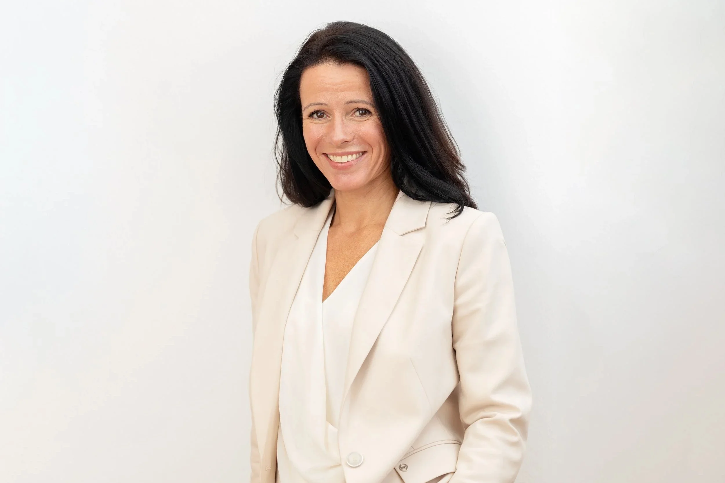 Portrait von Rosana Krönert – Frau mit dunklen, mittellangen Haaren, lachend, trägt eine cremefarbene Anzugjacke vor einem weißen Hintergrund.