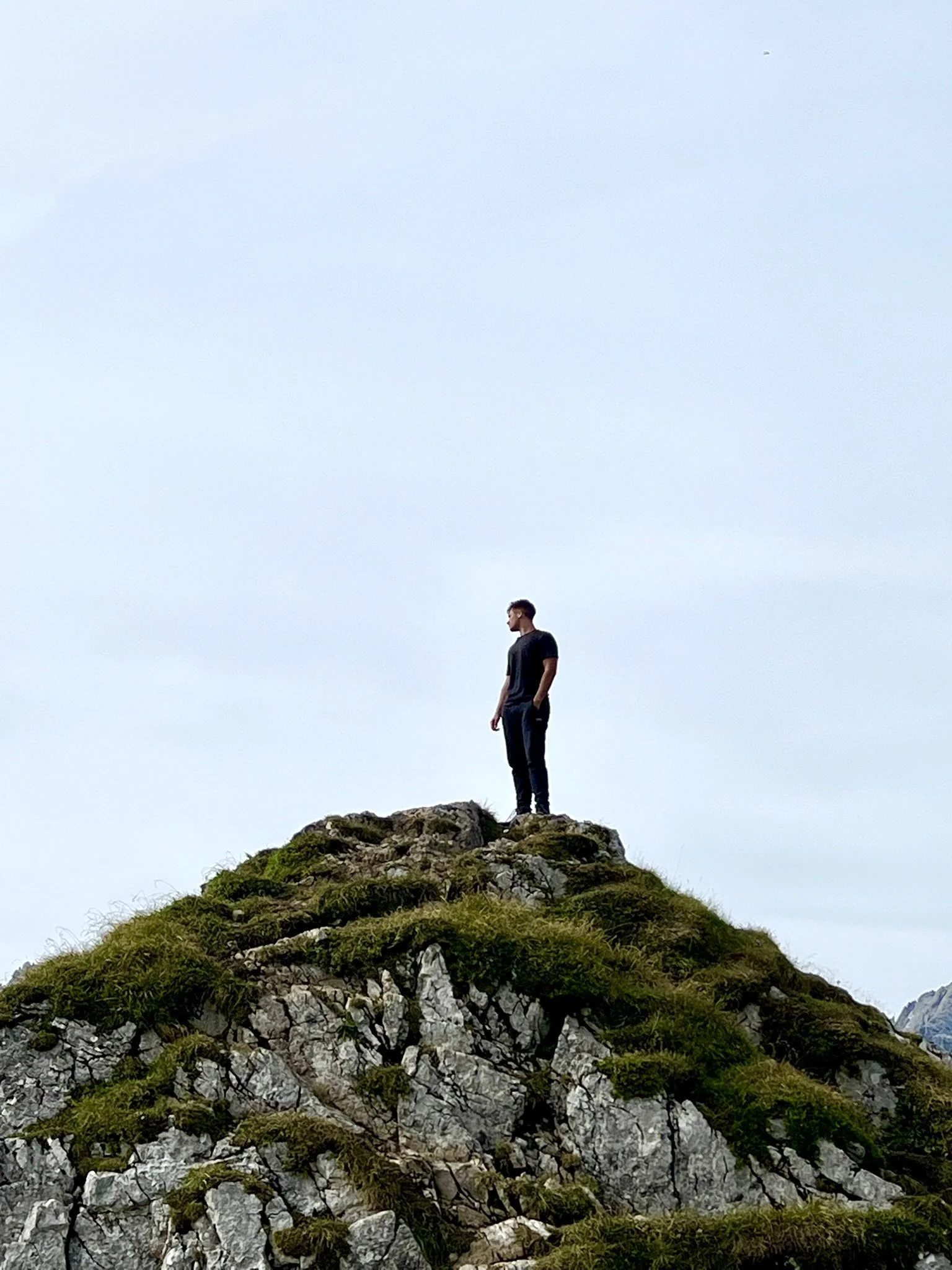 Ein Mann steht auf einem felsigen Hügel mit grüner Vegetation, blickt nach rechts, vor einem hellblauen Himmel.