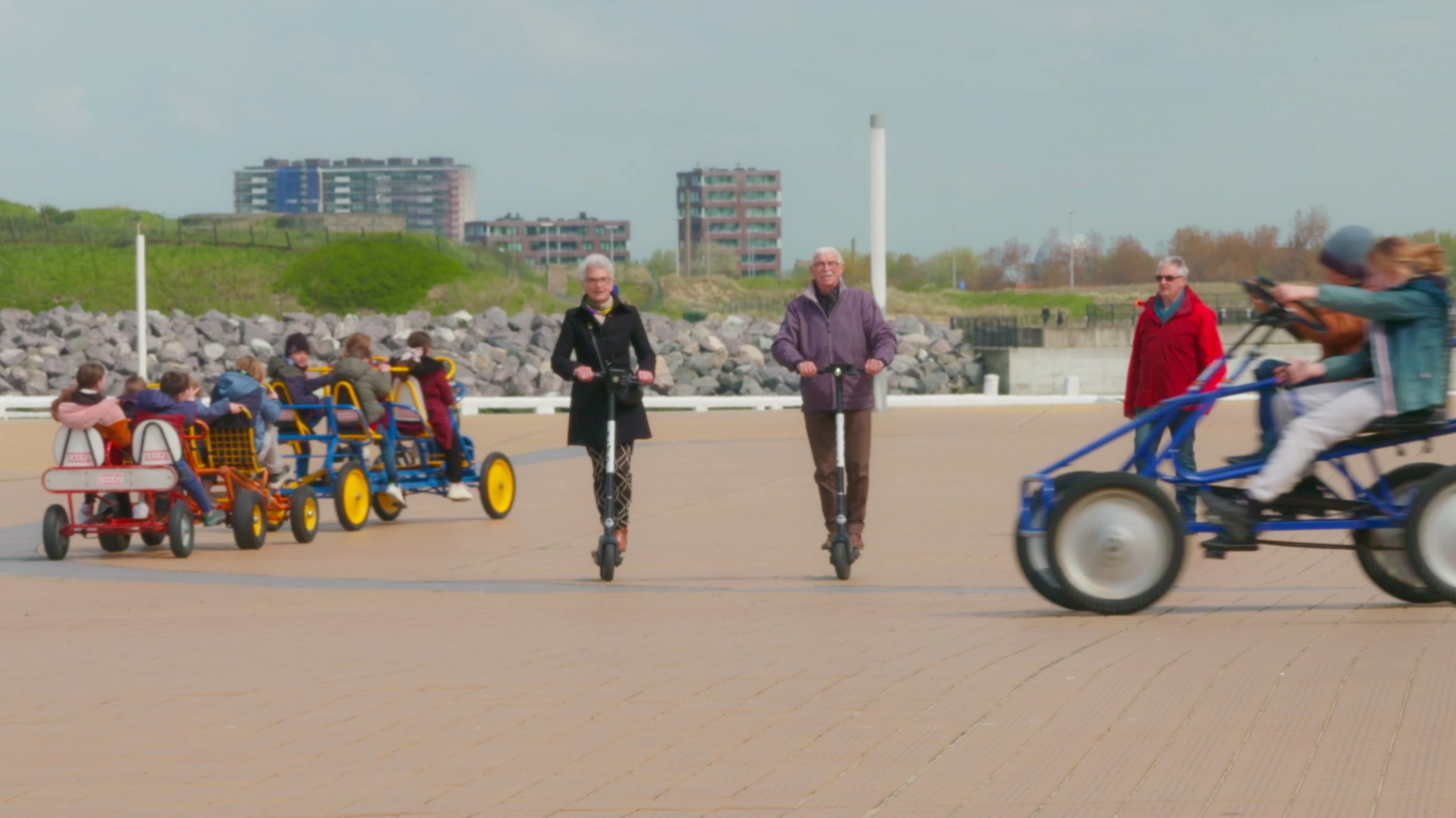 People riding pedal boats and scooters on a promenade near a rocky shoreline with buildings and greenery in the background.