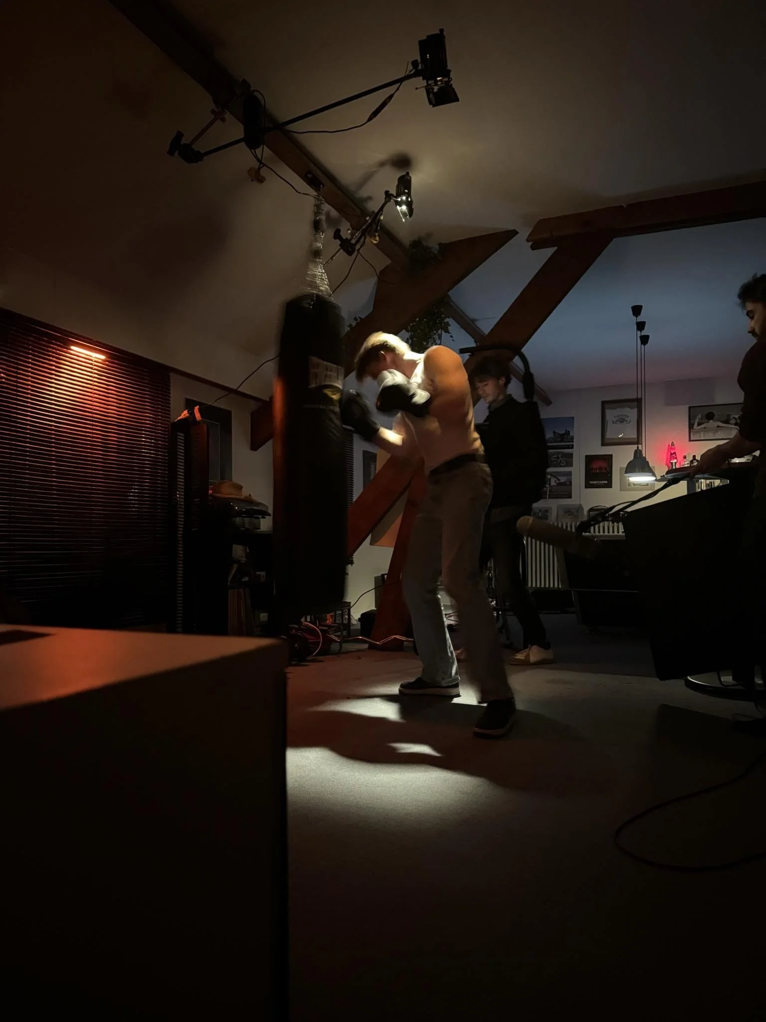 Person practicing boxing with a punching bag in a dimly lit room with other people and musical instruments in the background.