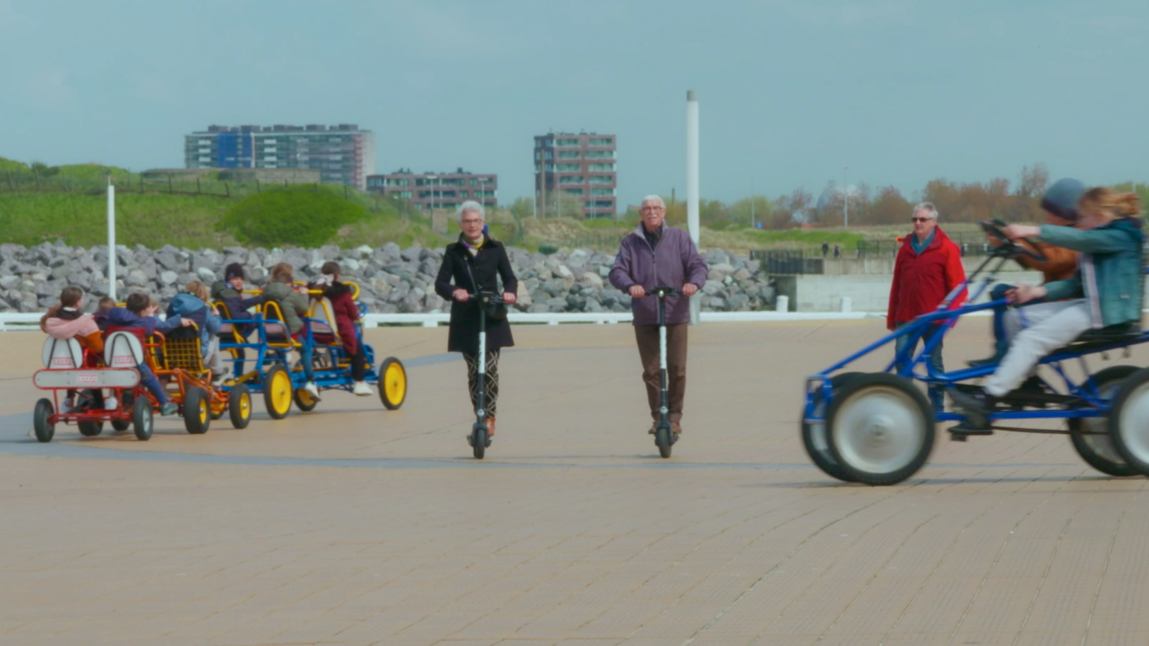 People riding on pedal-powered buggies along a promenade with green hills, buildings, and a cloudy sky in the background.