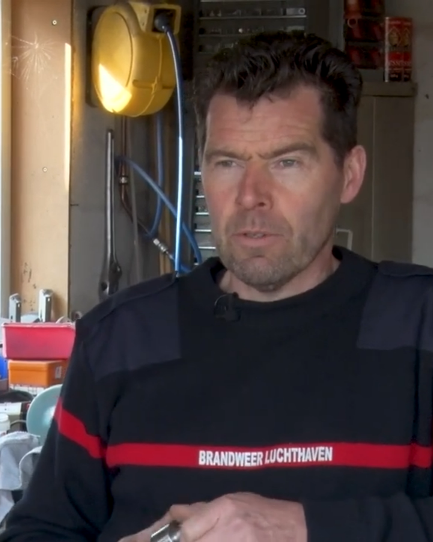 A man with dark hair and a beard wearing a black firefighter uniform with 'BRANDWEER LUCHTHAVEN' written across the chest, standing in a workshop or garage with tools and equipment around.
