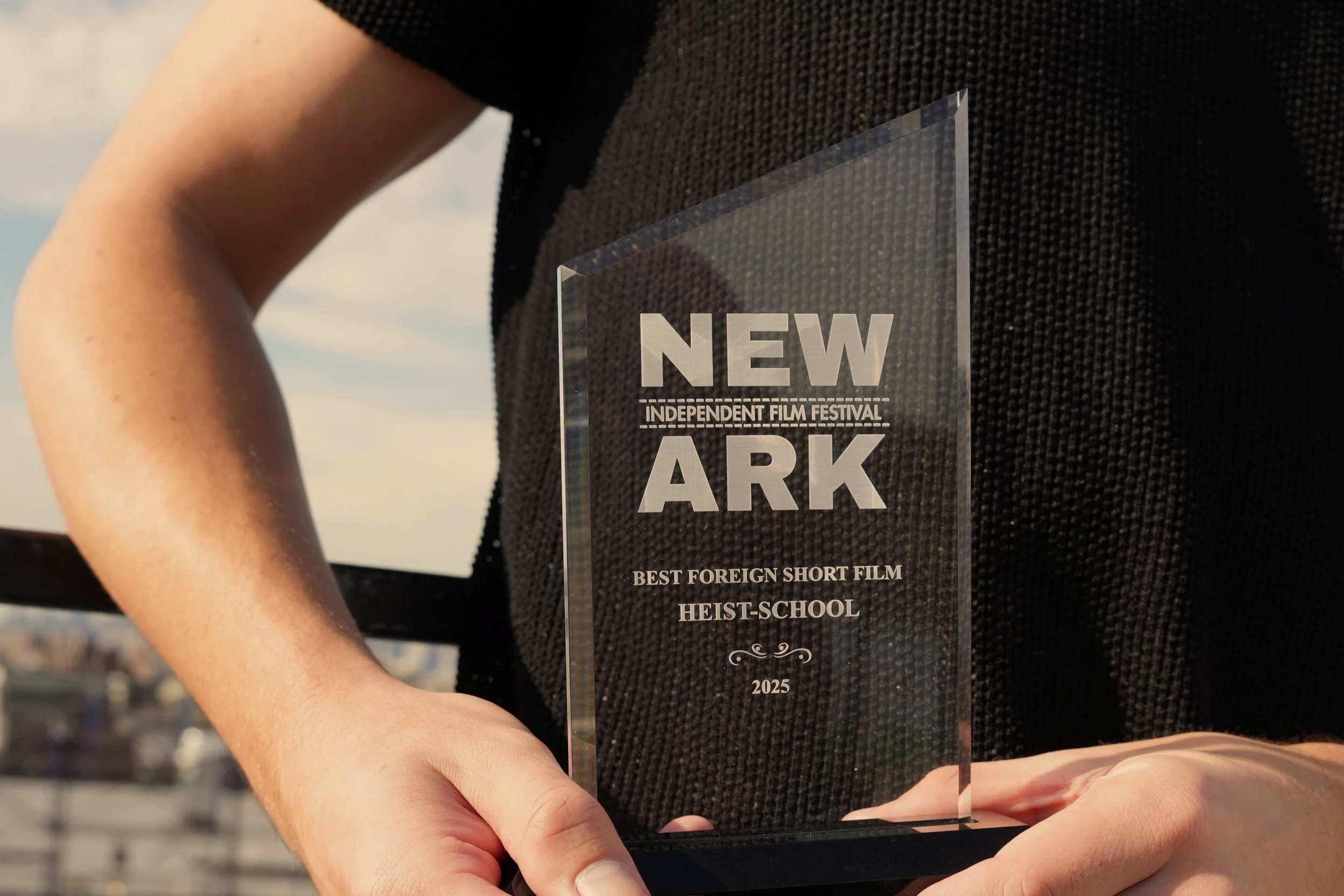 Person holding a clear glass award with the inscription 'NEWARK INDEPENDENT FILM FESTIVAL BEST FOREIGN SHORT FILM HEIST-SCHOOL 2025' against a cityscape background.