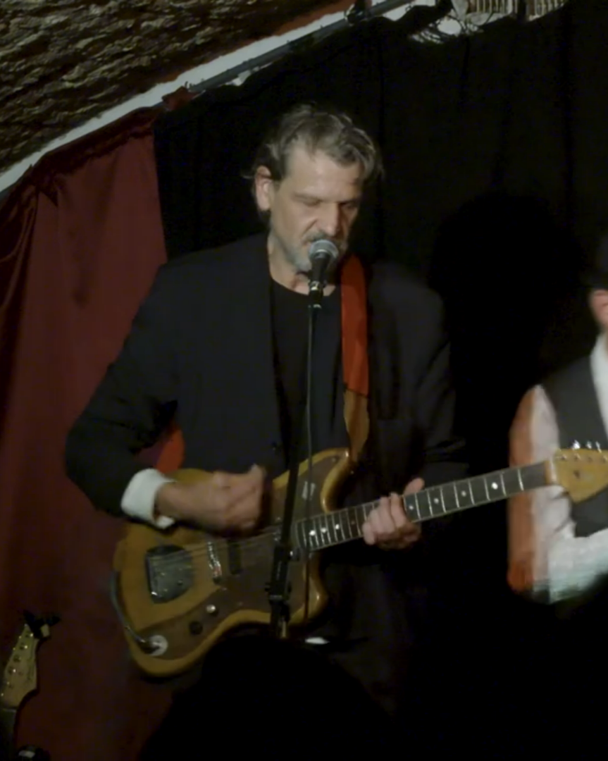 A middle-aged man with gray hair and a beard is performing on stage with a small electric guitar, singing into a microphone, in a dimly lit setting with a dark background and red curtain.