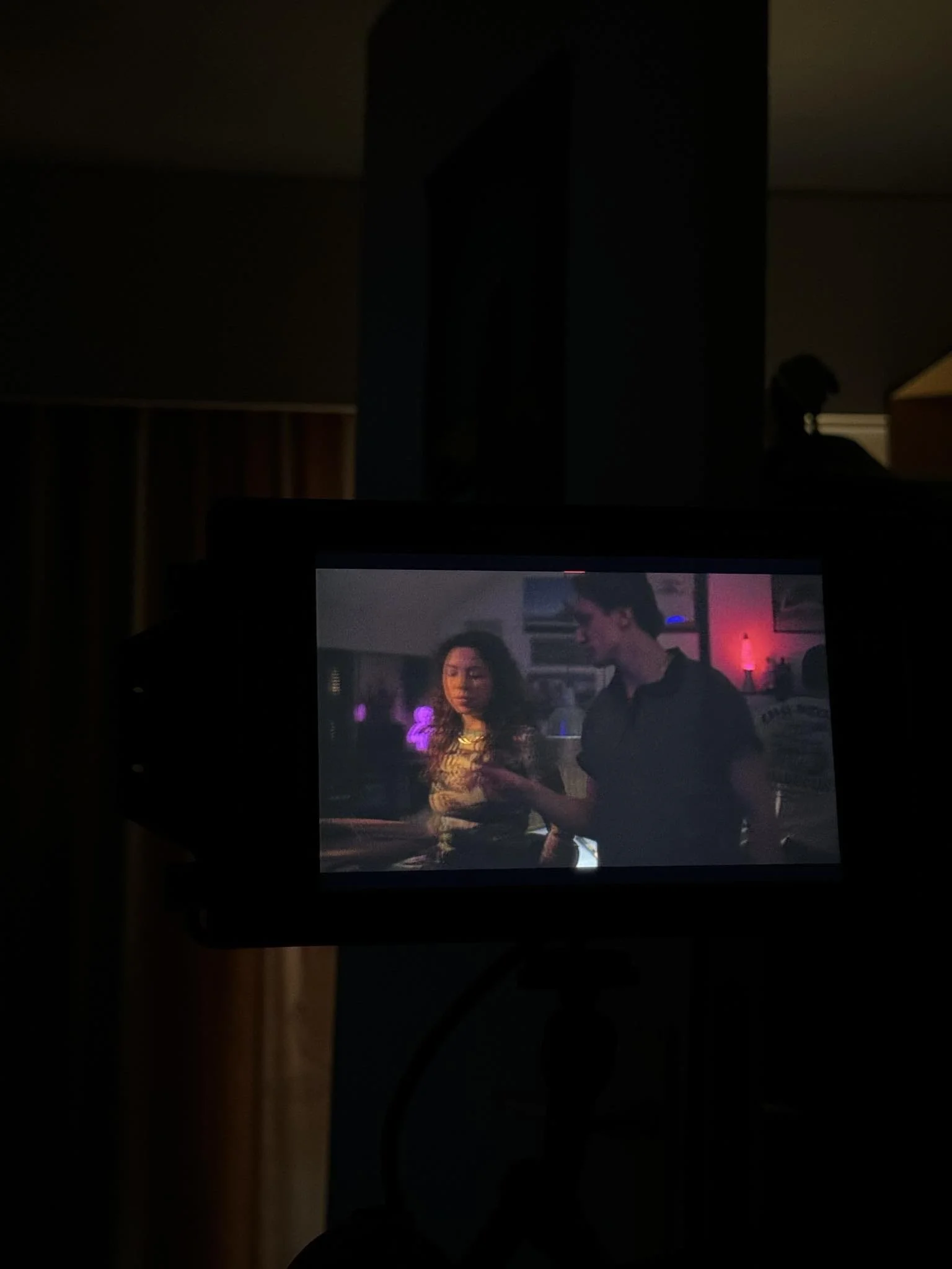 A camera monitor displaying a scene with a man and a woman in a dimly lit room, possibly a nightclub or bar, with colorful lighting and a relaxed atmosphere. The man is directing the actress.