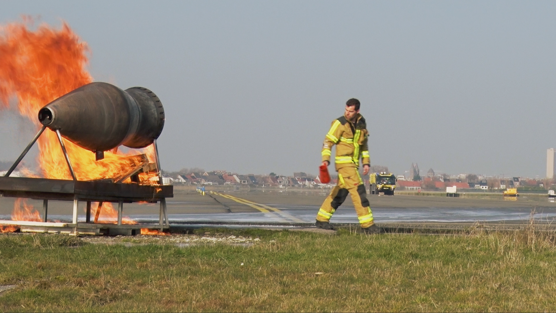 A firefighter inspecting a small fire on an runway, with flames emerging from a metal object mounted on a stand, during daytime.